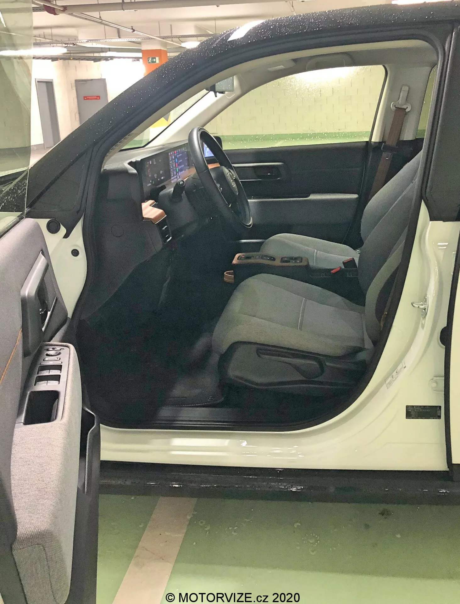 Vue rapprochée de la porte côté conducteur ouverte d'une voiture depuis un angle légèrement élevé, se concentrant sur l'intérieur moderne et axé sur la technologie. Un grand écran tactile central proéminent, des boutons physiques minimaux et des commandes montées sur le volant mettent en valeur les systèmes avancés d'infodivertissement et de contrôle. Le siège du conducteur suggère le confort avec un appuie-tête réglable. Les détails incluent des compartiments de rangement dans la porte, une grille de haut-parleur, des commandes de vitres électriques, des ceintures de sécurité, des leviers de réglage et des tapis de sol, signalant un modèle plus récent et bien entretenu.