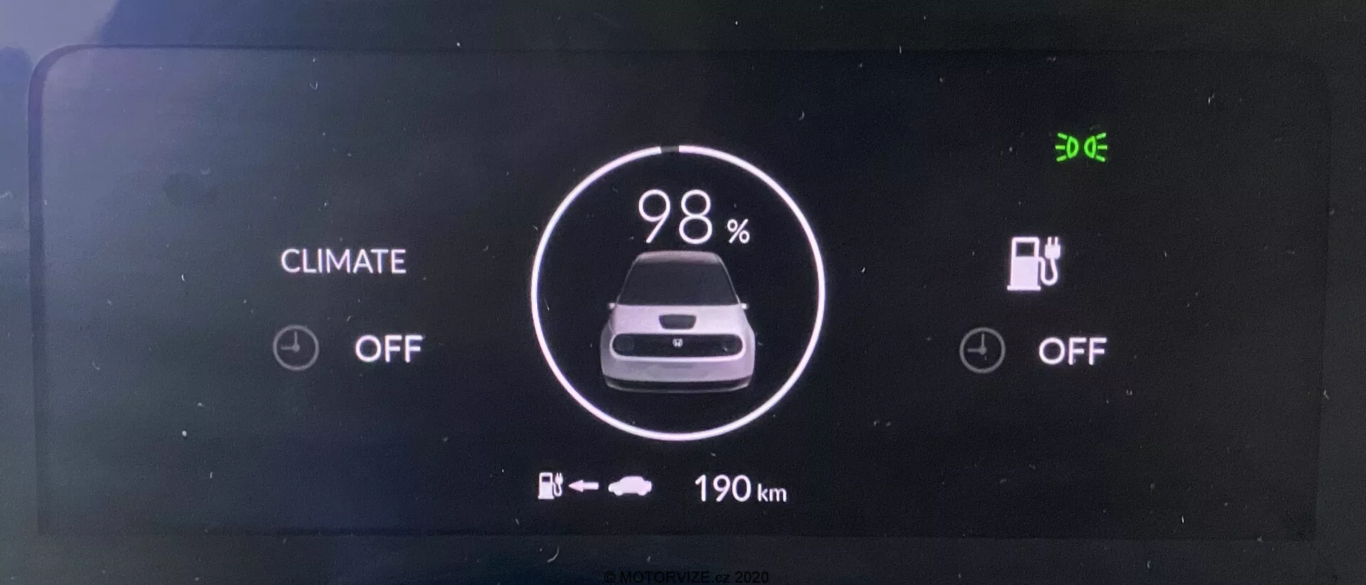 Tableau de bord numérique du véhicule électrique Honda e 2020. L'écran, vu de face, affiche clairement un indicateur de batterie à 98% de charge, une icône de voiture et '190 km' d'autonomie restante en dessous. Les systèmes de contrôle du climat et de charge sont tous deux 'OFF'. L'heure '9:06' est affichée dans le coin supérieur gauche.