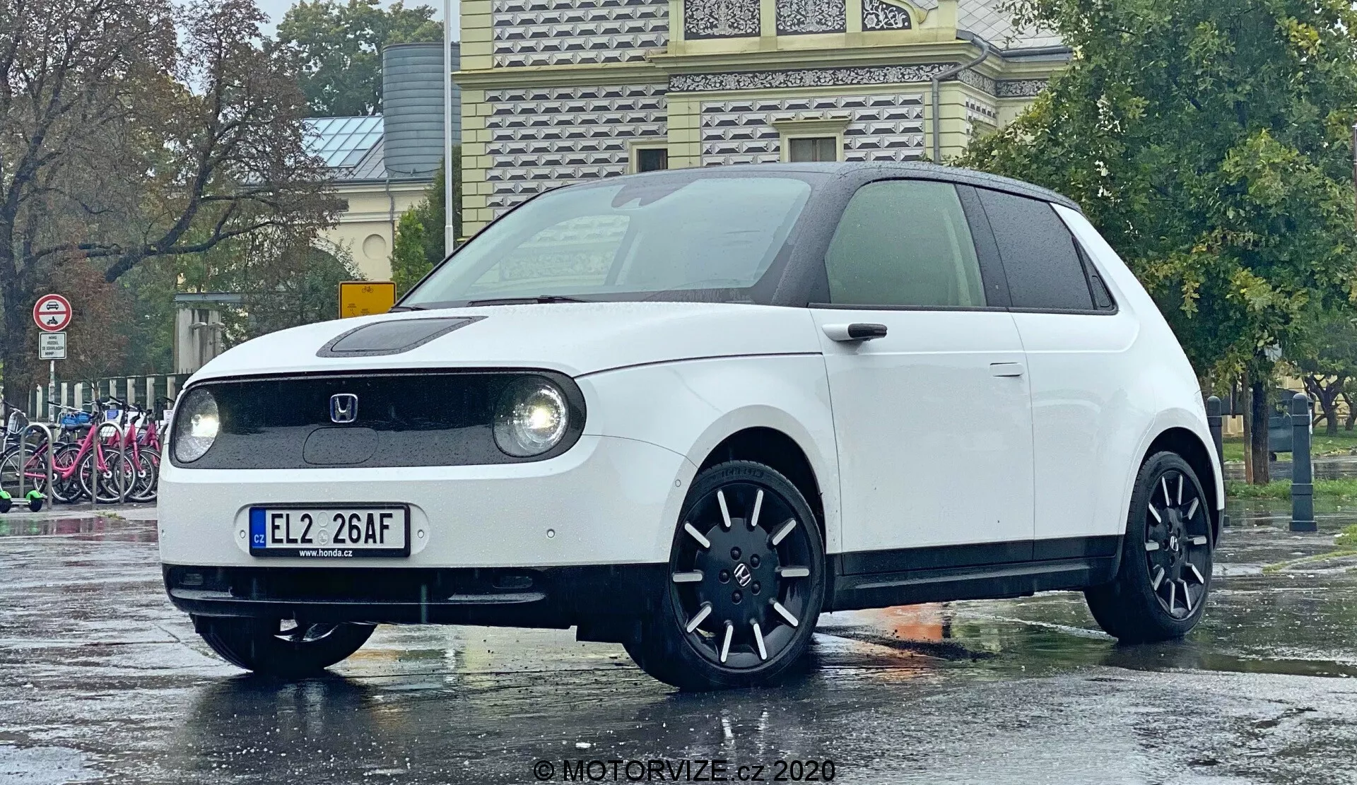 Honda e 2020 photographiée en contre-plongée accentuant son avant et son profil latéral partiel, mettant en avant les phares LED circulaires dans des boîtiers rectangulaires, une calandre avant fermée typique des véhicules électriques, un badge de fabricant placé au centre, des jantes en alliage foncé à multiples rayons et un design minimaliste rétro-moderne. La compacte hatchback est sur un sol mouillé reflétant un environnement pluvieux, ce qui rend ses lignes nettes et sa couleur plus vives.