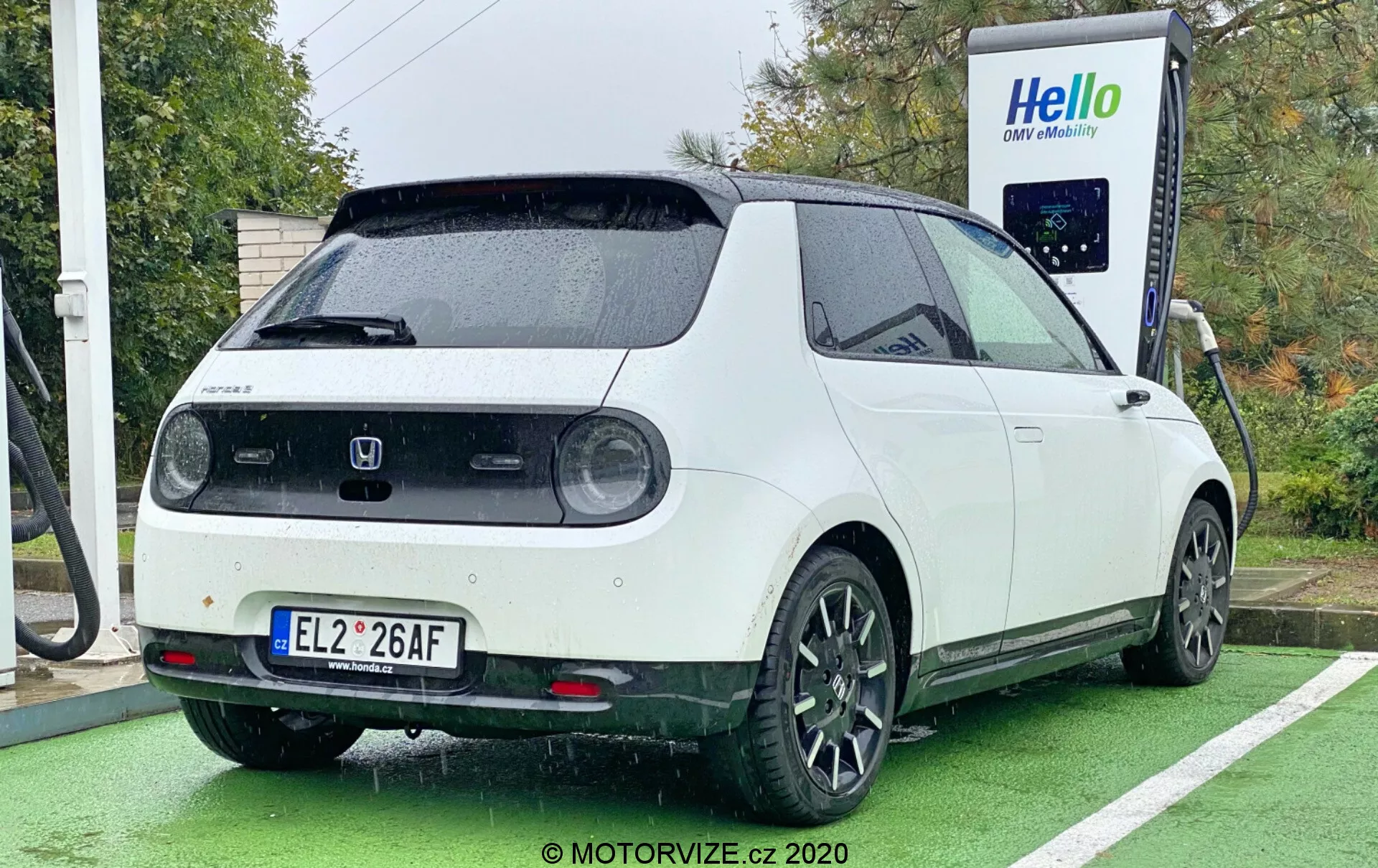 Un véhicule électrique Honda e 2020 photographié sous un angle arrière trois-quarts, montrant le côté arrière droit, la face latérale partielle et le hayon arrière. La voiture, avec une combinaison de couleurs bicolore avec une carrosserie blanche et un toit noir, est garée à une station de recharge avec le câble de recharge connecté au port situé sur le côté arrière. Le design du véhicule inclut des feux arrière circulaires, un grand essuie-glace arrière, des capteurs de stationnement, un spoiler de toit et des jantes en alliage noir à cinq branches. L'image met également en avant l'esthétique rétro-moderne et carrée de la voiture, prise en journée par temps nuageux ou pluvieux, avec le véhicule garé dans un emplacement spécialement désigné pour la recharge des voitures électriques, peint en vert.