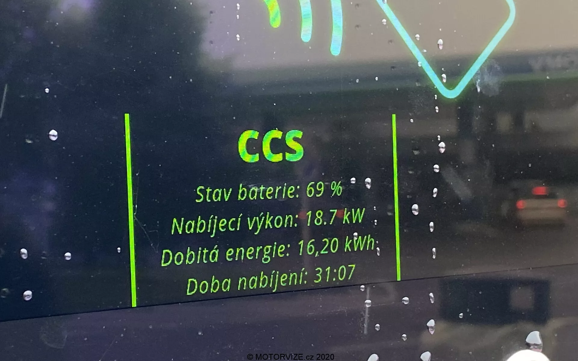 Vue intérieure à travers le pare-brise d'un véhicule électrique, se concentrant sur le reflet de l'interface de la station de recharge sur le verre. Les détails clés incluent 'CCS' pour Combined Charging System, 'État de la batterie : 69 %,' 'Puissance de charge : 18,7 kW,' 'Énergie rechargée: 16,20 kWh,' et 'Temps de charge: 31:07.' Le reflet apparaît fantomatique sur un fond pluvieux avec une autre voiture visible au loin.
