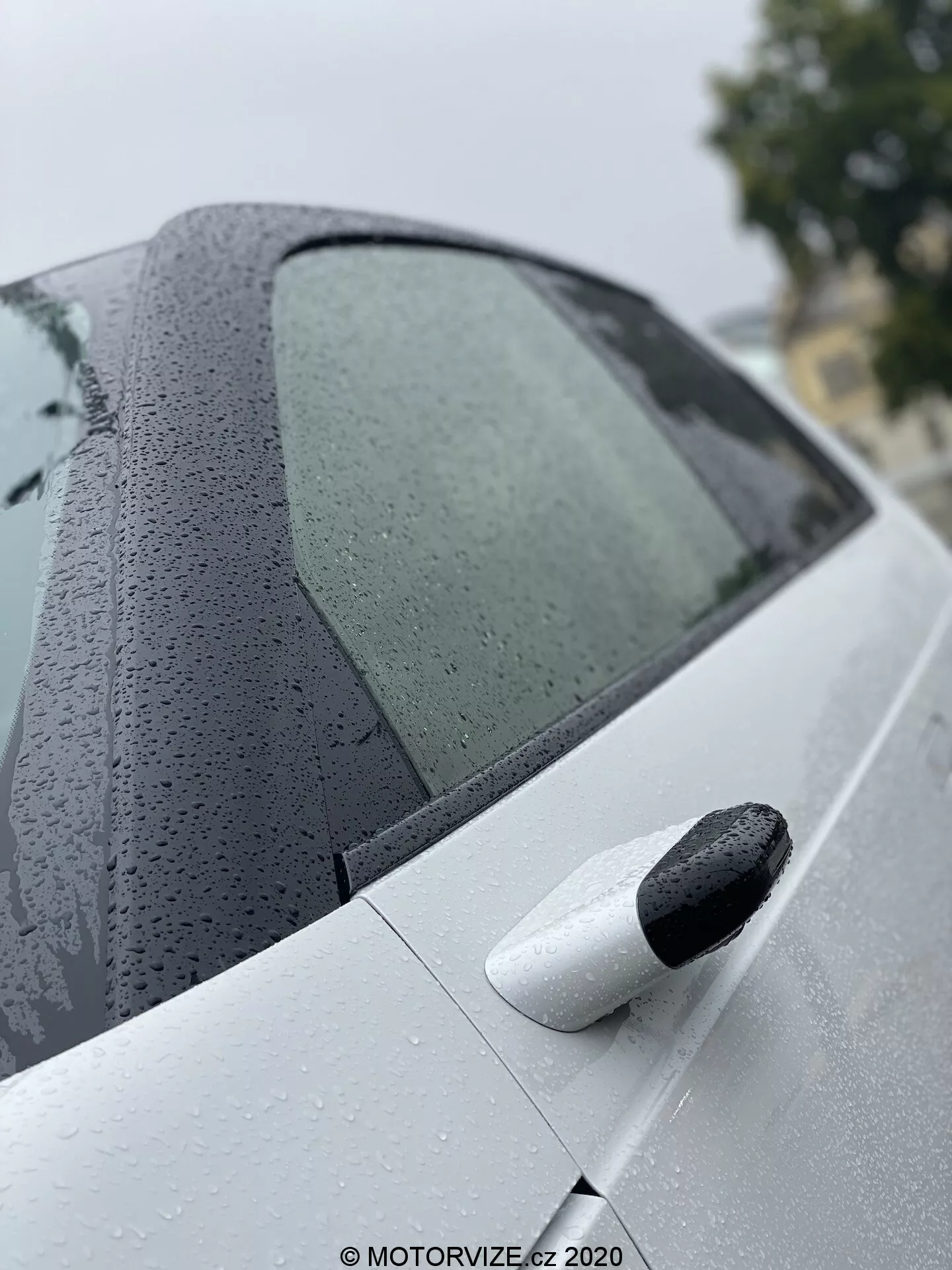 Vue rapprochée et angulaire de la section arrière de la Honda e 2020, mettant en avant le design de la fenêtre latérale passager arrière avec des gouttes d'eau, et la porte arrière de couleur claire avec une poignée de porte noire, soulignant des caractéristiques de revêtement de peinture avancé et un système d'entrée sans clé.