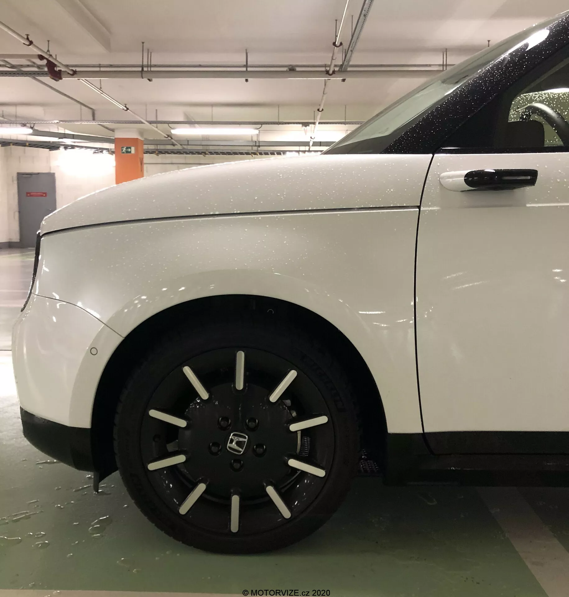 Vue latérale rapprochée de la roue avant et du quart de panneau de la Honda E 2020 blanche, mettant en valeur la jante en alliage à dix rayons et la carrosserie propre et aérodynamique. L'image, probablement prise dans un parking couvert, met en évidence l'excellent état du véhicule et son design robuste, avec des surfaces mouillées visibles indiquant une exposition récente à l'eau.