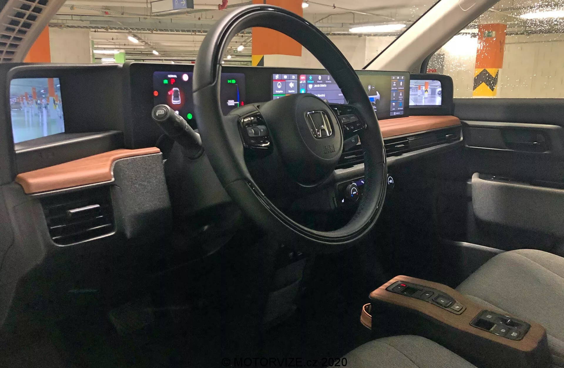 Vue intérieure de la Honda e 2020, centrée sur la zone du conducteur vue du centre du véhicule. Le tableau de bord présente un design moderne avec un schéma de couleurs bicolores. Le volant, avec le logo Honda et plusieurs boutons de commande, est au milieu, entouré d'un panneau d'affichage numérique à la place des jauges analogiques. À droite, la console centrale numérique comprend des panneaux tactiles pour l'audio, la navigation, le contrôle de la climatisation et d'autres réglages. Le design met l'accent sur l'intégration technologique, minimisant les boutons physiques en faveur des interfaces tactiles. La voiture semble être stationnée dans un garage, avec des lumières fluorescentes et des piliers en béton visibles à travers les fenêtres.