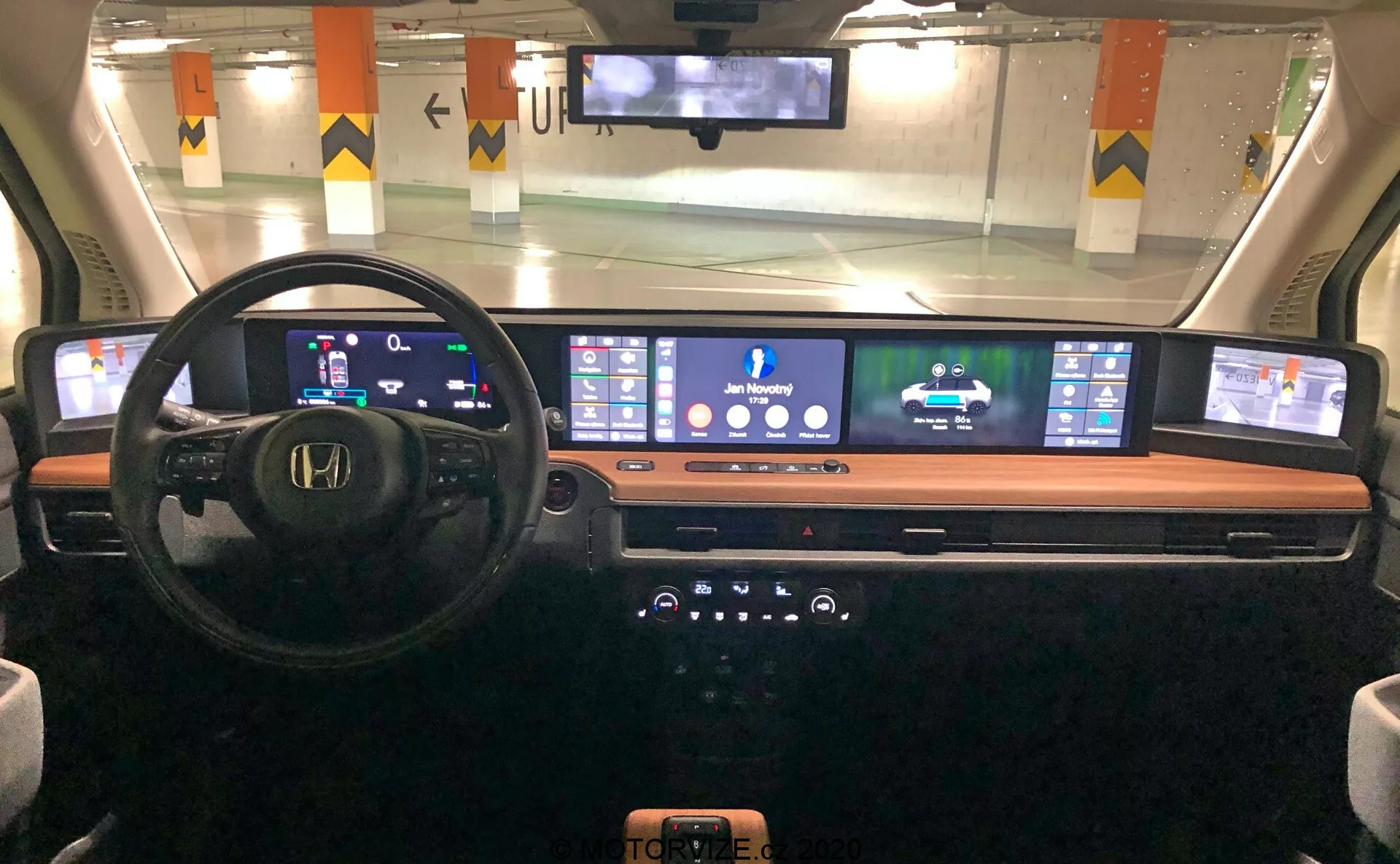 Vue intérieure du tableau de bord de la Honda e 2020 depuis la perspective du conducteur, affichant un tableau de bord numérique, volant multifonction, écran numérique continu expansif pour la navigation et les médias, console centrale élégante avec boutons et boutons, et un chargeur de téléphone sans fil, mettant en valeur la technologie automobile moderne et le design.