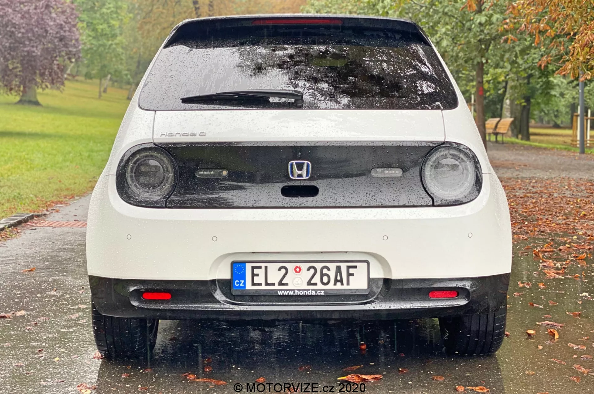 Vue arrière d'une Honda e 2020 blanche et mouillée, affichant des feux arrière à LED circulaires, un troisième feu stop, un emblème Honda, un hayon arrière avec essuie-glace, une plaque d'immatriculation européenne, des capteurs de stationnement, des feux antibrouillard arrière, un spoiler arrière et des éléments réfléchissants sur le pare-chocs. L'image capture des détails fins comme les gouttes d'eau sur le véhicule, avec un arrière-plan flou de parc avec des arbres et de l'herbe.