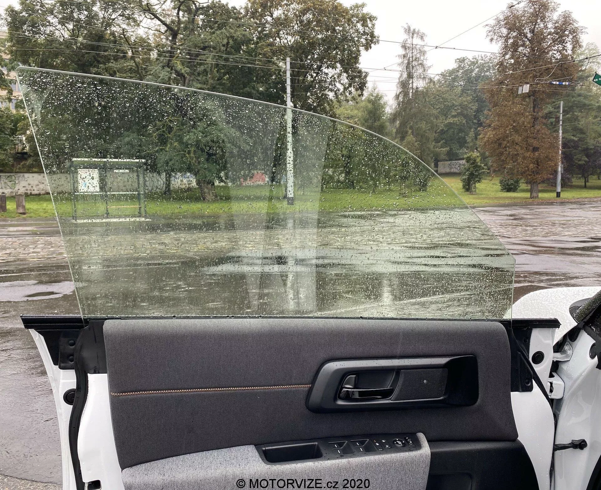 Vue intérieure depuis la perspective du passager dans une Honda e 2020, montrant le panneau de porte droit avec insert en tissu et poignée métallique, le pare-brise avec des gouttes de pluie et une vue sur une route mouillée avec des arbres et une clôture au-delà.