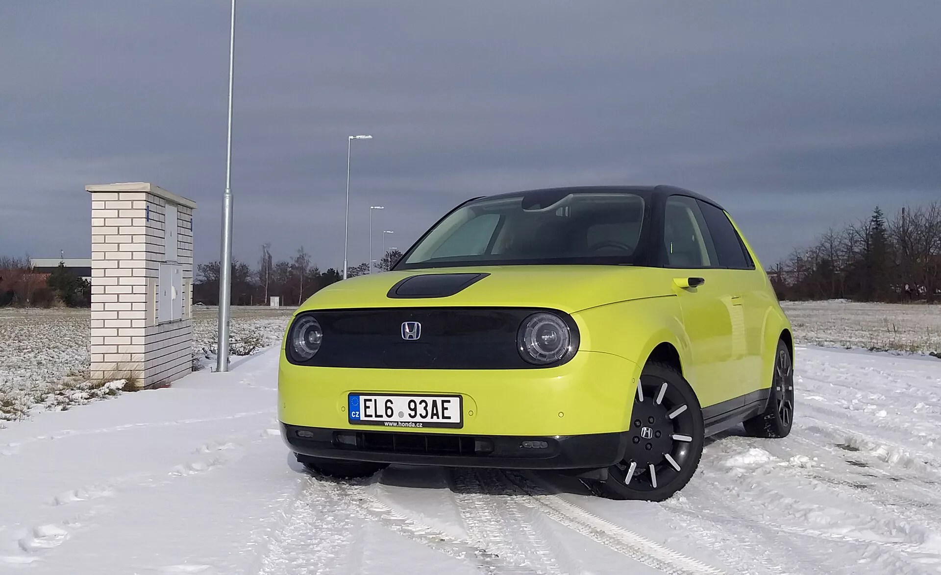 Honda e 2020 en jaune vif sur une surface couverte de neige avec ciel nuageux, comportant des phares LED, une technologie de véhicule électrique avec assistance avancée au conducteur, des jantes en alliage noir et des pneus hiver.