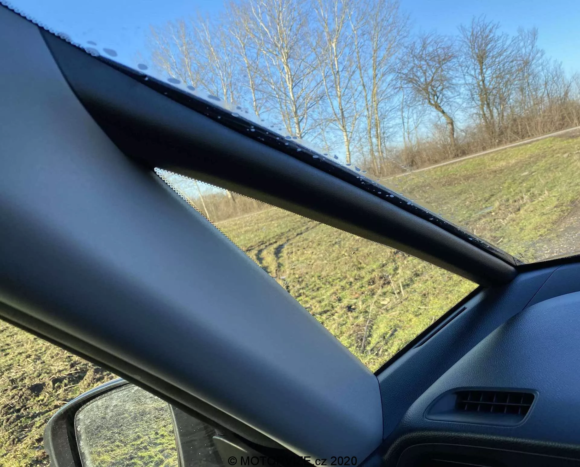 Vue intérieure d'une Honda Jazz Crosstar 2020 regardant vers la vitre côté conducteur, centrée sur le pilier A. Le pilier A est recouvert d'un matériau texturé non métallique et obscurcit partiellement la vue. La vitre latérale montre des gouttes d'eau, indiquant une pluie récente ou de la condensation, avec une zone herbeuse et des arbres visibles à l'extérieur sous un ciel bleu clair.
