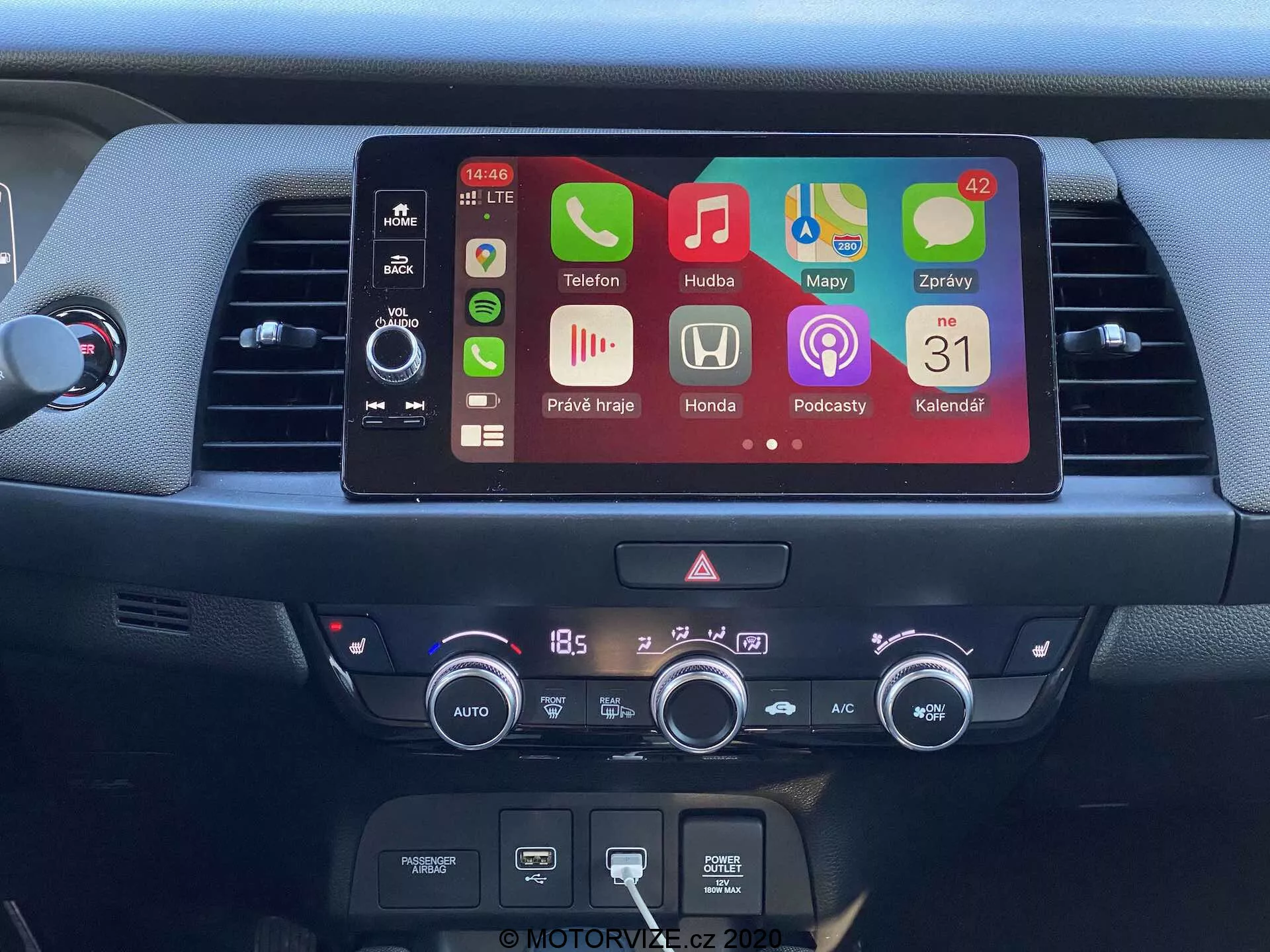 Vue détaillée de la console centrale de la Honda Jazz 2020, mettant en avant un écran tactile d'infodivertissement moderne avec des icônes d'applications colorées incluant téléphone, musique, navigation, réglages de la voiture, podcasts et calendrier. Sous l'écran se trouvent des commandes physiques du système audio, une fente pour lecteur CD et un système de contrôle climatique numérique réglé à 18,5 degrés Celsius. L'image met en avant des fonctionnalités avancées telles que la connectivité Bluetooth, l'intégration des smartphones et le contrôle climatique automatique.