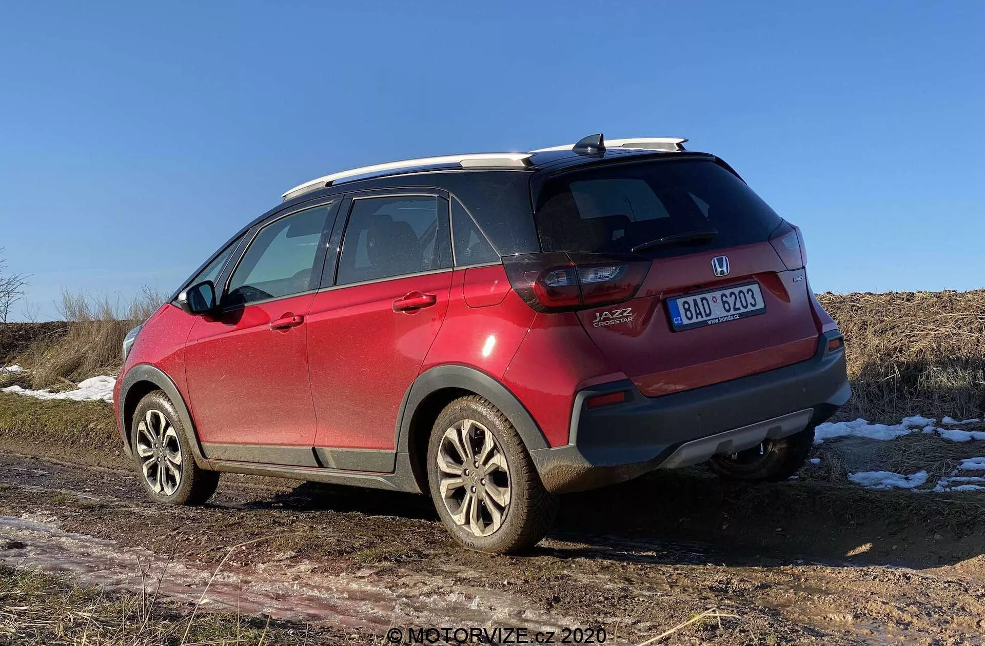 Un SUV Honda Jazz Crosstar rouge de 2020 vu de trois quarts arrière, montrant des détails tels que les badges de marque et de modèle, les feux arrière à LED, le pare-chocs arrière avec des rayures et des capteurs, les jantes en alliage, les pneus toutes saisons, les barres de toit, l'antenne aileron de requin, les vitres teintées avec des lignes de dégivrage et une plaque d'immatriculation centrée. L'ouverture du coffre de type hayon et les plaques de neige au sol avec un champ sec en arrière-plan suggèrent une fin d'automne, un hiver ou un début de printemps par un ciel dégagé de midi ou début d'après-midi.