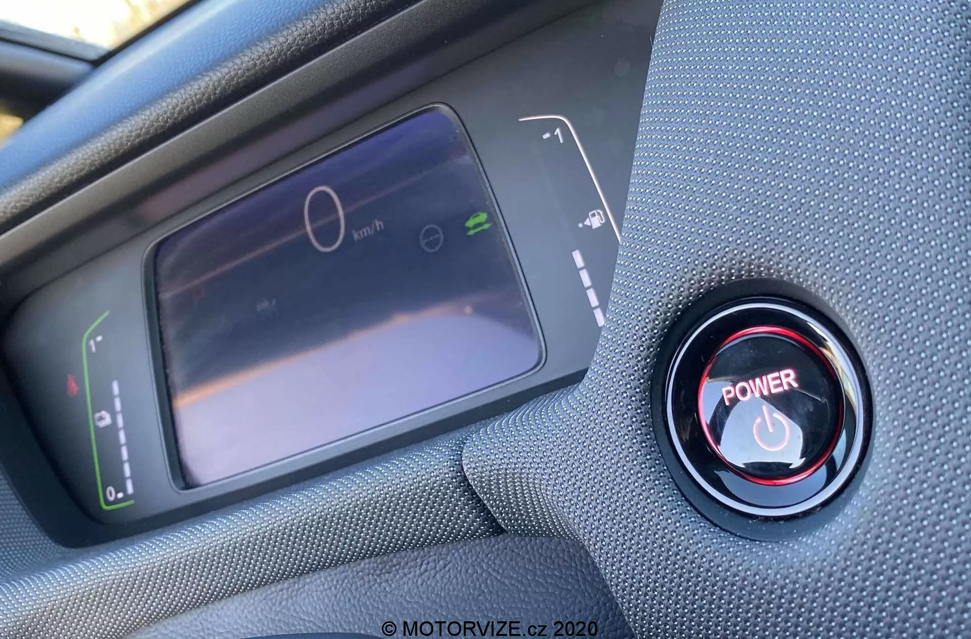 Vue intérieure en gros plan depuis la perspective du conducteur d'une Honda Jazz Crosstar 2020, se concentrant sur l'affichage numérique du tableau de bord et le bouton de démarrage/arrêt rouge du moteur. L'écran LCD à gauche affiche un compteur de vitesse à l'arrêt et un graphique de consommation d'énergie, tandis qu'à droite, le bouton de démarrage/arrêt rouge est entouré d'un anneau métallique. La photo est prise sous un angle oblique depuis le dessus, avec une mise au point nette sur le bouton et une mise au point douce sur l'affichage.