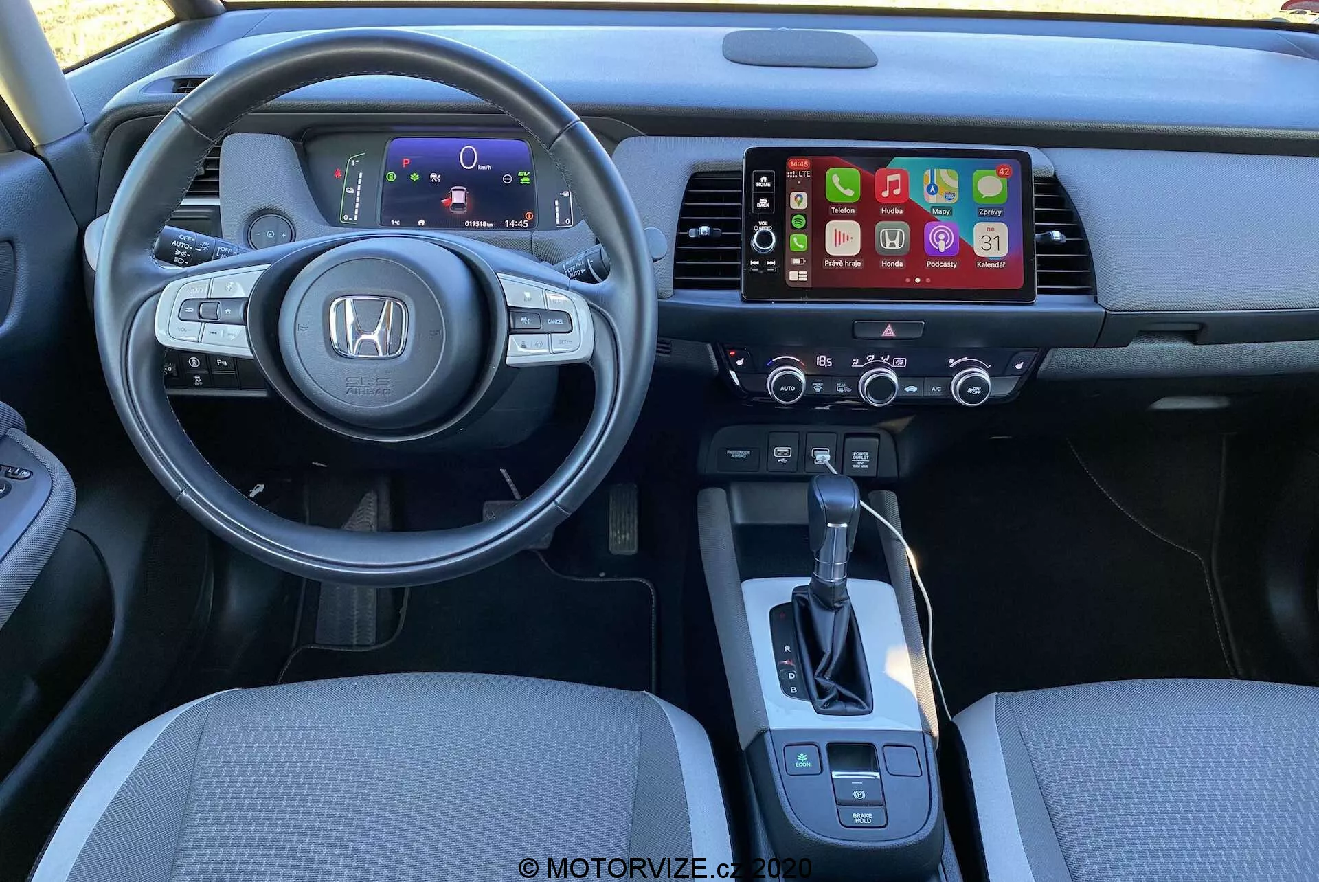 Vue intérieure de la Honda Jazz Crosstar 2020 depuis la perspective du conducteur. Un volant multifonctionnel moderne avec le logo Honda est au centre, doté de commandes pour l'audio, le régulateur de vitesse et les commandes vocales. Derrière le volant, un tableau de bord numérique affiche des informations essentielles. La console centrale abrite un système d'infodivertissement prenant probablement en charge Apple CarPlay ou Android Auto, avec des commandes de climat en dessous. Le levier de vitesse est simple, avec des commandes supplémentaires pour le frein de stationnement électronique et la sélection du mode de conduite. L'éclairage et l'angle mettent en avant le confort et la disposition ergonomique.