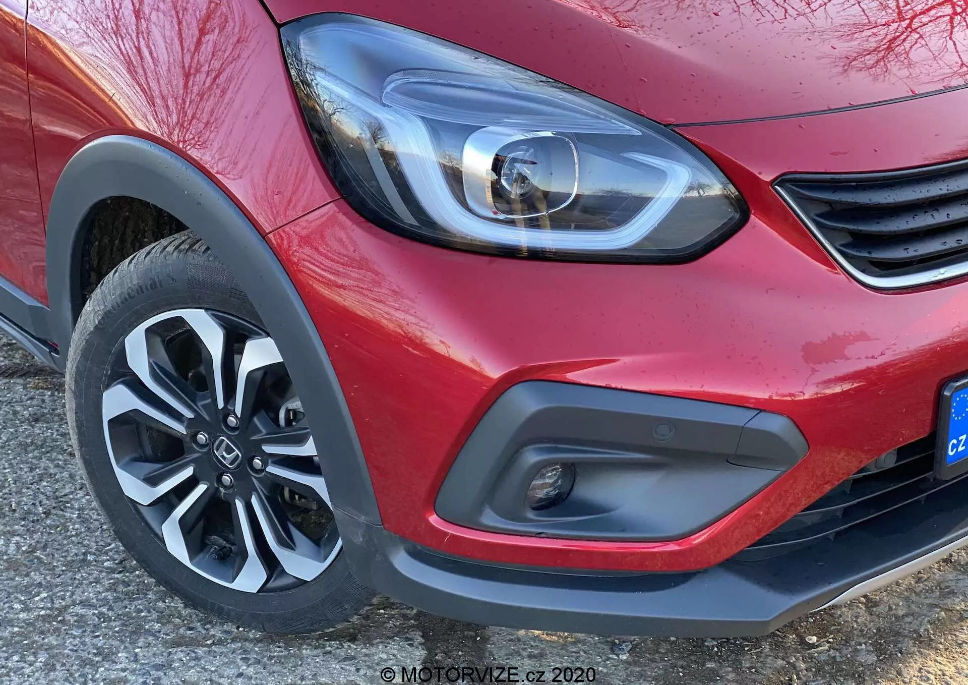 Vue en gros plan du coin avant d'une Honda Jazz 2020 (Jazz Crosstar) rouge, en se concentrant sur le phare LED moderne, le détail minimaliste de la calandre, le pare-chocs avant bicolore avec feu antibrouillard et la roue en alliage à cinq branches dynamique.