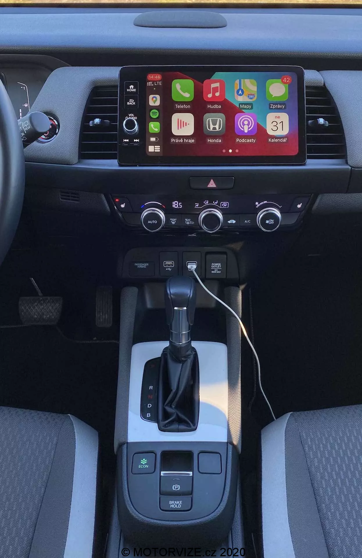 Vue intérieure de la console centrale de la Honda Jazz Crosstar 2020 sous un angle légèrement surélevé depuis l'arrière. Se concentre sur la zone du levier de vitesses, le sélecteur de transmission automatique et l'écran tactile d'infodivertissement affichant plusieurs icônes d'applications, suggérant des options de connectivité avancées comme Apple CarPlay et Android Auto. Sous l'écran, il y a des bouches d'aération et un panneau de commande de climatisation bizone avec des boutons et des cadrans physiques. Le levier de vitesses est flanqué du sélecteur de mode de conduite et des boutons de frein de stationnement électronique, avec un espace de rangement ouvert à droite indiquant des ports de charge. Les sièges avant bicolores mettent en valeur le design intérieur et le confort.