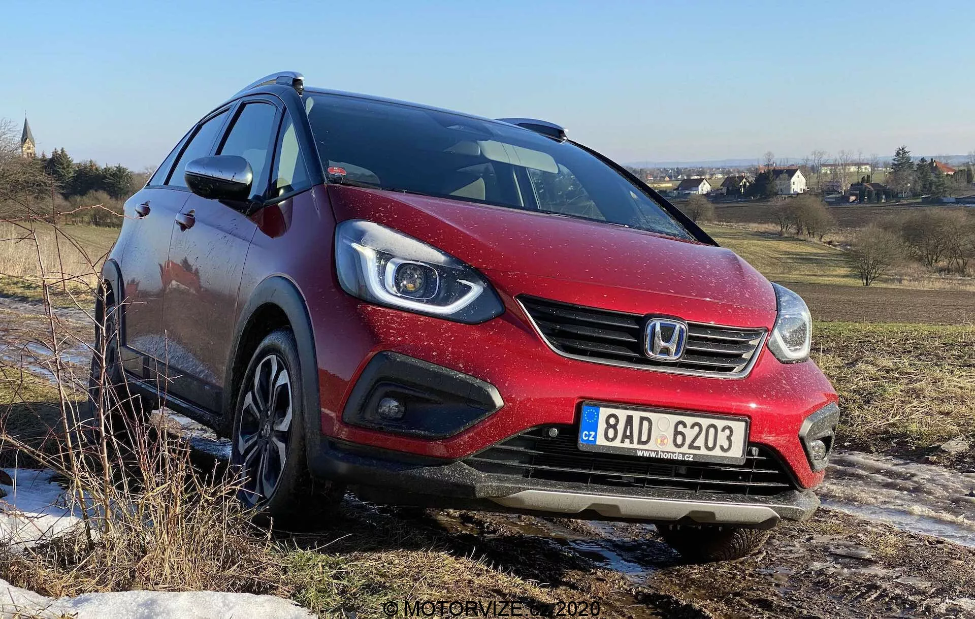 Vue de trois quarts avant d'une Honda Jazz Crosstar 2020 rouge, prise en contre-plongée, montrant sa calandre avant, ses phares halogènes à projecteur avec feux de jour à LED intégrés, la calandre avant avec le logo de la marque et les roues en alliage. La voiture est sur un terrain accidenté sous un ciel bleu clair, suggérant sa capacité à gérer des surfaces non pavées.