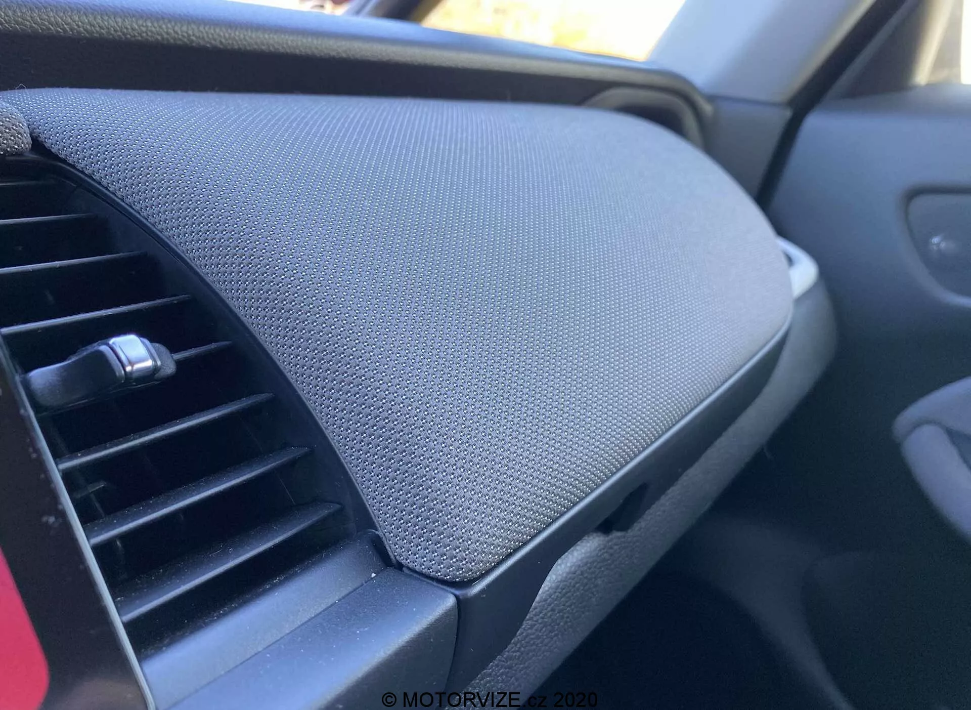 Vue rapprochée de l'intérieur d'un véhicule Honda Jazz 2020 (Jazz Crosstar), centré sur le tableau de bord. Pris de la perspective du passager, l'image montre la bouche d'aération à gauche, avec un design réglable horizontal. Dominant la partie droite de l'image se trouve le compartiment de rangement du tableau de bord avec un couvercle fermé, caractérisé par une surface texturée, possiblement en maille ou en tissu. Le compartiment semble intégré sans poignée visible, suggérant un mécanisme d'ouverture par pression. Le schéma de couleurs global est sombre, impliquant une esthétique neutre visant à réduire les distractions.