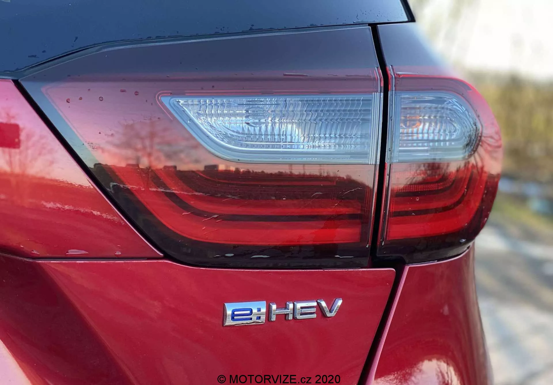 Photo détaillée en contre-plongée de la section arrière d'une Honda Jazz Crosstar 2020 capturant le couvercle du coffre, le feu arrière à LED moderne et l'insigne du véhicule hybride électrique e:HEV. Met en valeur la finition peinture métallisée rouge brillant et la qualité de construction supérieure avec des joints de panneau étroits, indiqués par des lignes propres et des reflets à la lumière naturelle.