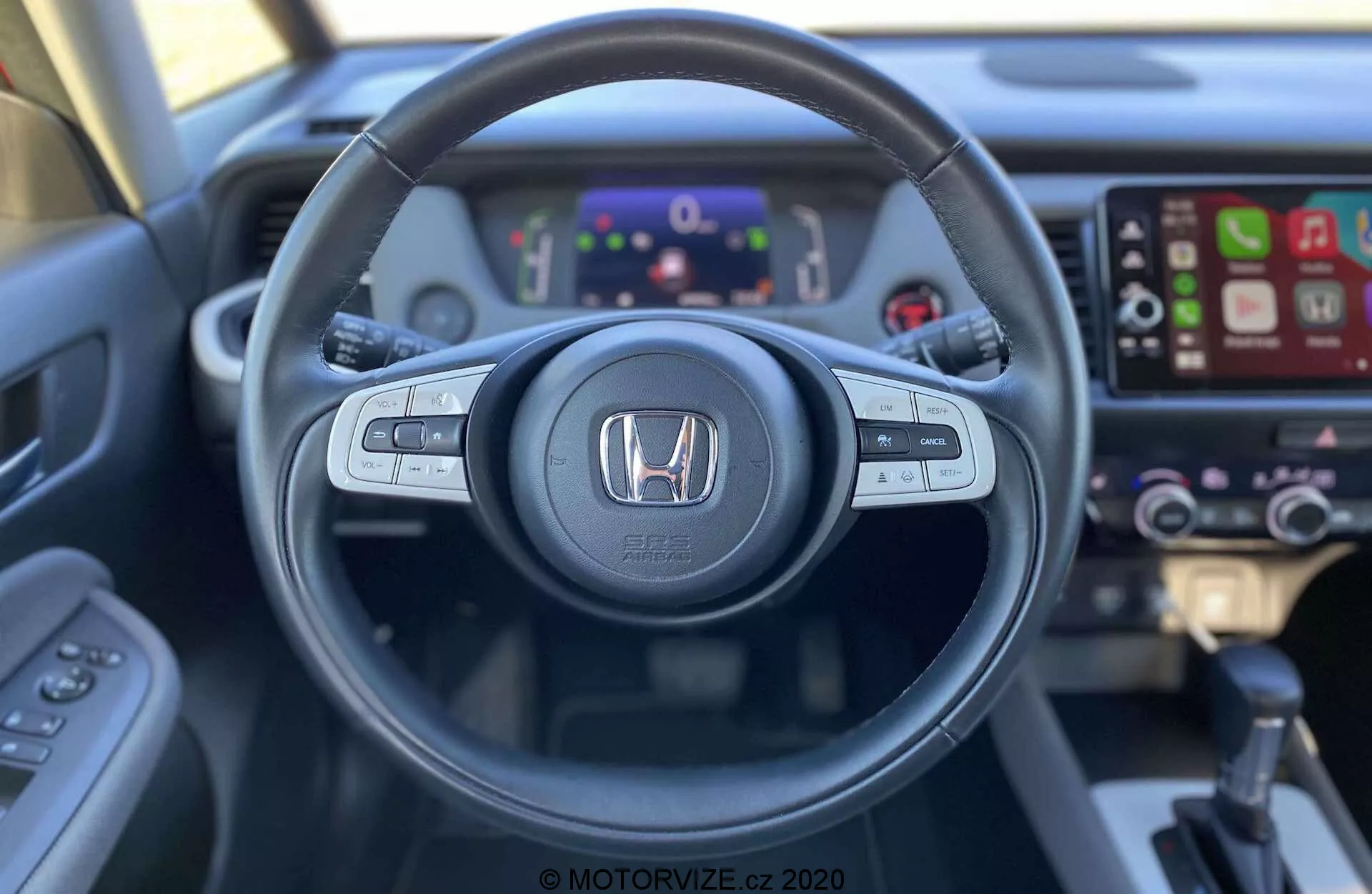 Vue intérieure de la Honda Jazz Crosstar 2020 depuis le siège du conducteur, avec un volant équipé de commandes audio, de contrôle vocal et de régulateur de vitesse. Le logo Honda est bien visible au centre. Derrière le volant, un tableau de bord numérique affiche divers indicateurs et informations. À droite, un système d'infodivertissement à écran tactile compatible avec Apple CarPlay est visible, ainsi que des commandes de climatisation légèrement floues. L'image, prise d'une perspective à la première personne, met en avant le design ergonomique et l'accessibilité de la technologie du véhicule.
