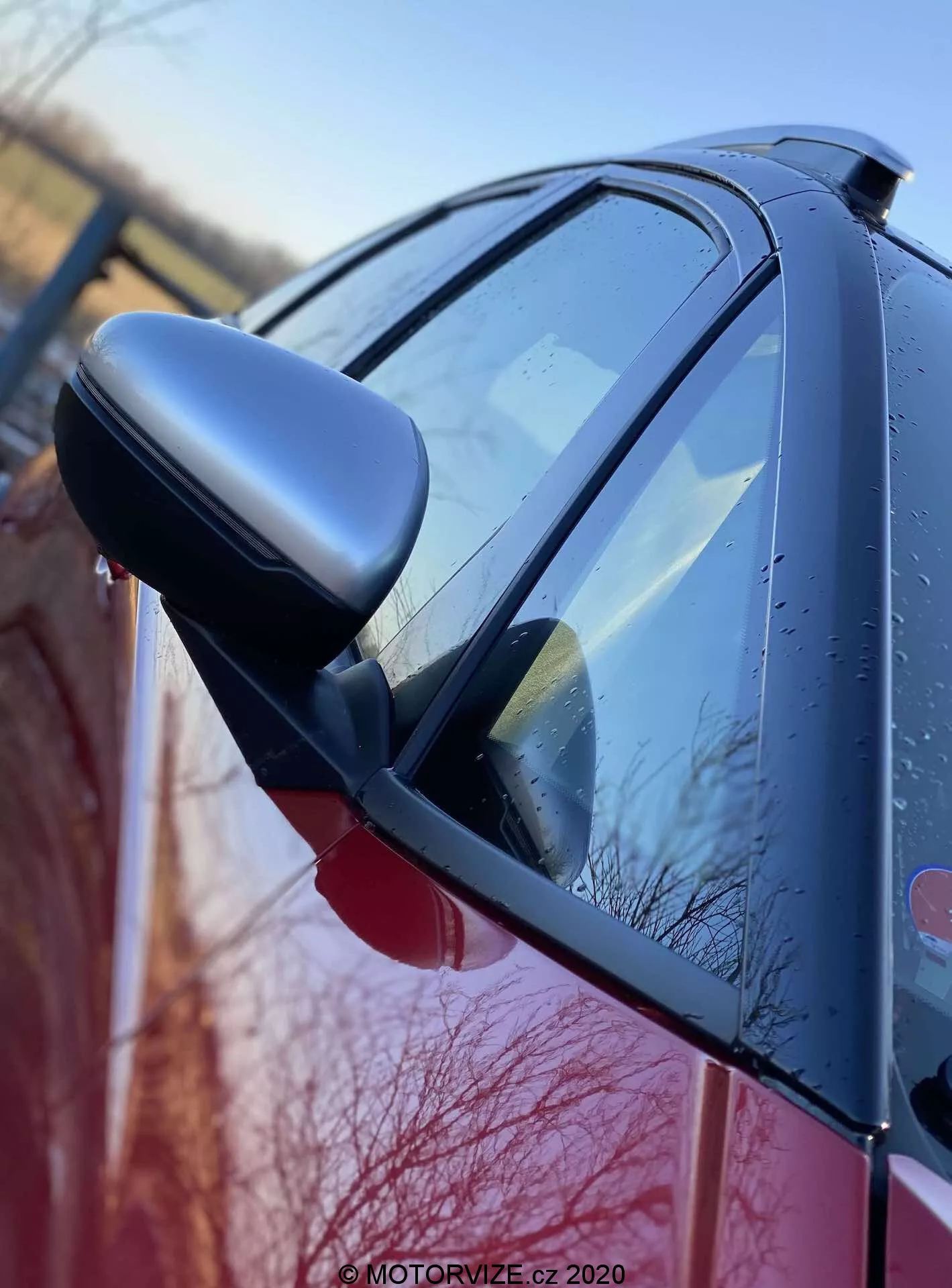 Vue latérale rapprochée de la Honda Jazz 2020 (Jazz Crosstar) mettant en avant le rétroviseur côté conducteur avec des indicateurs de direction intégrés. L'image, prise sous un angle bas près de l'avant de la voiture, capture la carrosserie brillante, possiblement rouge, avec des gouttes d'eau, suggérant une pluie récente ou une rosée du matin. Le miroir à coque foncée et le design aérodynamique de la voiture sont mis en évidence, soulignant la finition et l'état extérieur.