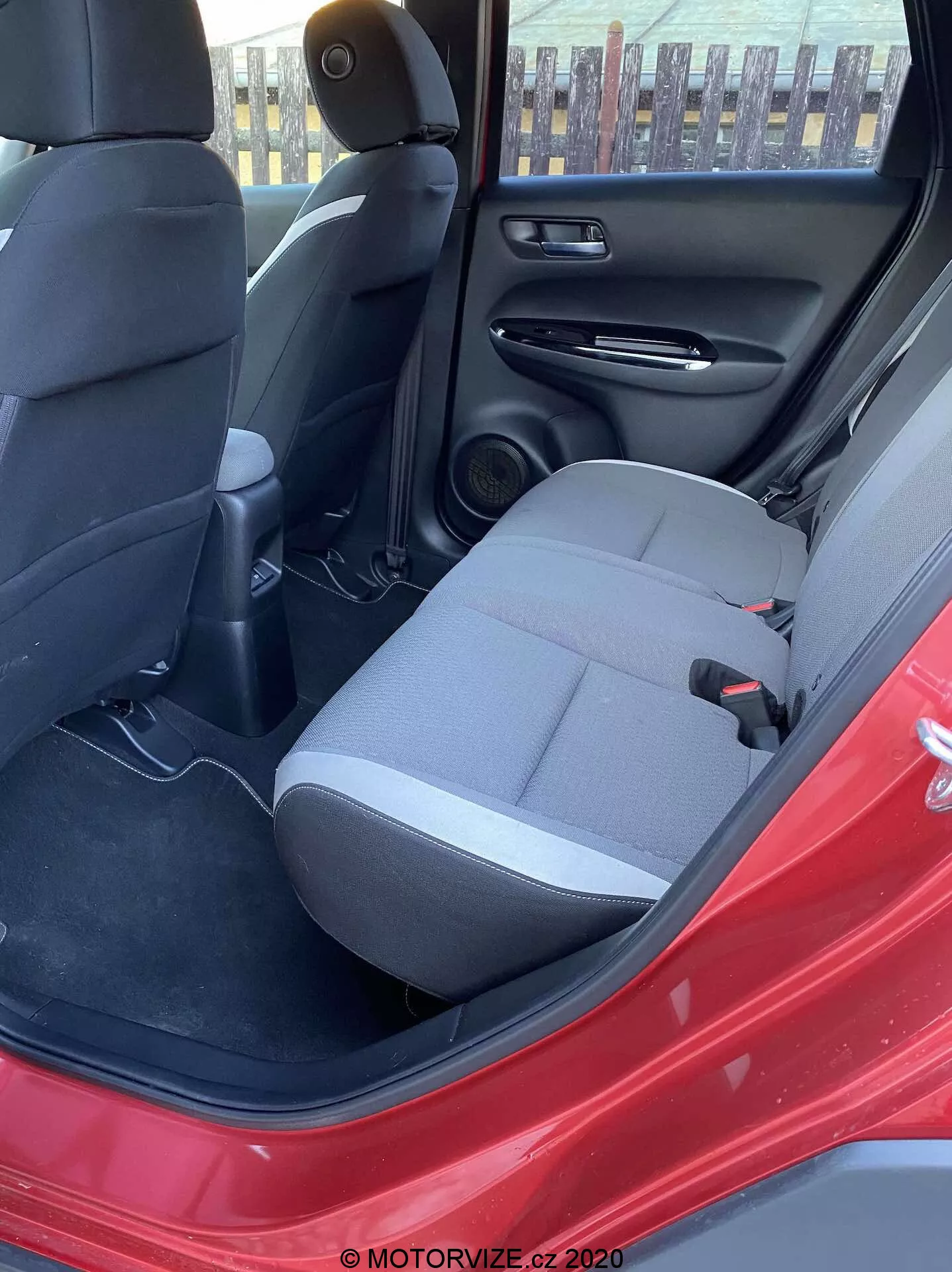 Vue intérieure de la Honda Jazz Crosstar 2020 depuis la porte arrière du côté conducteur ouverte, mettant en valeur le siège passager arrière en tissu foncé, la ceinture de sécurité, la console centrale arrière avec des bouches d'aération, le haut-parleur dans la porte arrière, le panneau de porte intérieur noir avec une poche de rangement, le revêtement de sol en moquette et la couleur extérieure rouge éclatante de la voiture. La lumière naturelle du jour illumine la scène.