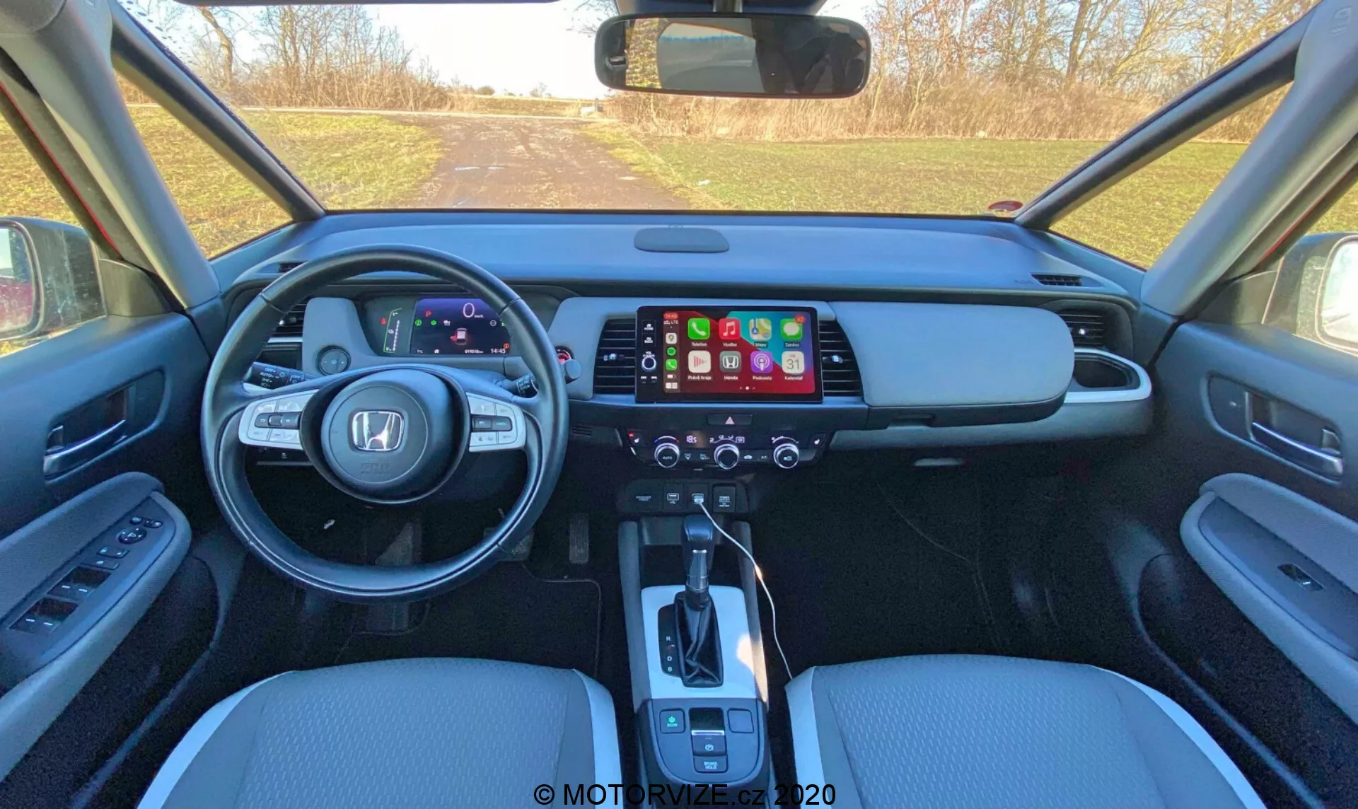 Vue intérieure de la Honda Jazz 2020 (Jazz Crosstar) observée depuis la zone des sièges arrière, présentant le tableau de bord avant avec un tableau de bord numérique, un grand écran tactile d'infodivertissement, un panneau de contrôle de la climatisation, un levier de changement de vitesse automatique, des sièges en cuir et des commandes de panneau de porte.