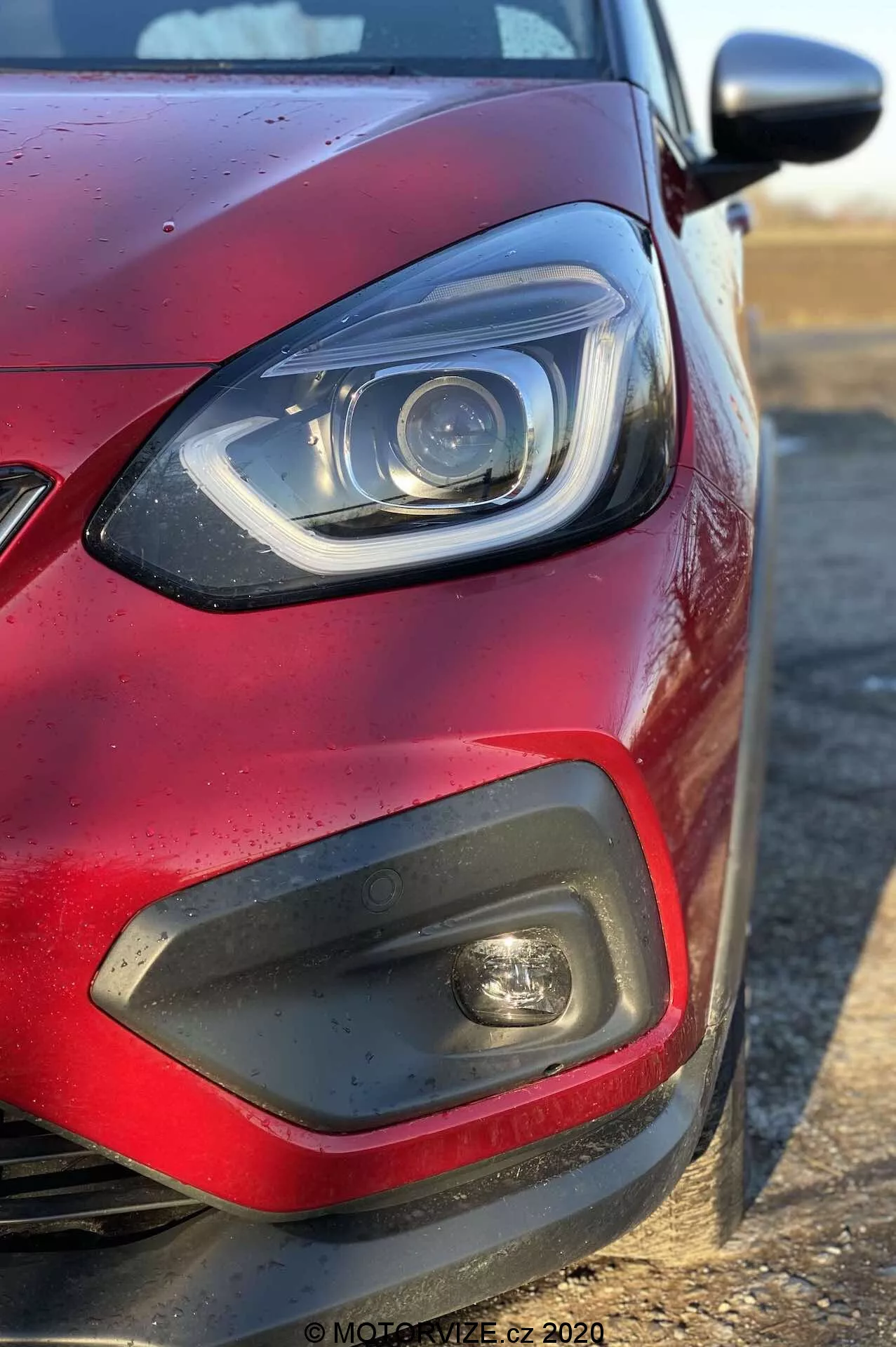 Gros plan du quart avant-gauche d'une Honda Jazz 2020 (Jazz-Crosstar) rouge sous un angle latéral, se concentrant sur le phare avec éléments LED ou xénon et une barre DRL incurvée, un insert en plastique noir avec un feu antibrouillard, une partie de la calandre, une peinture rouge métallisée avec des gouttes d'eau et un rétroviseur latéral de la couleur de la carrosserie s'étendant depuis la droite.