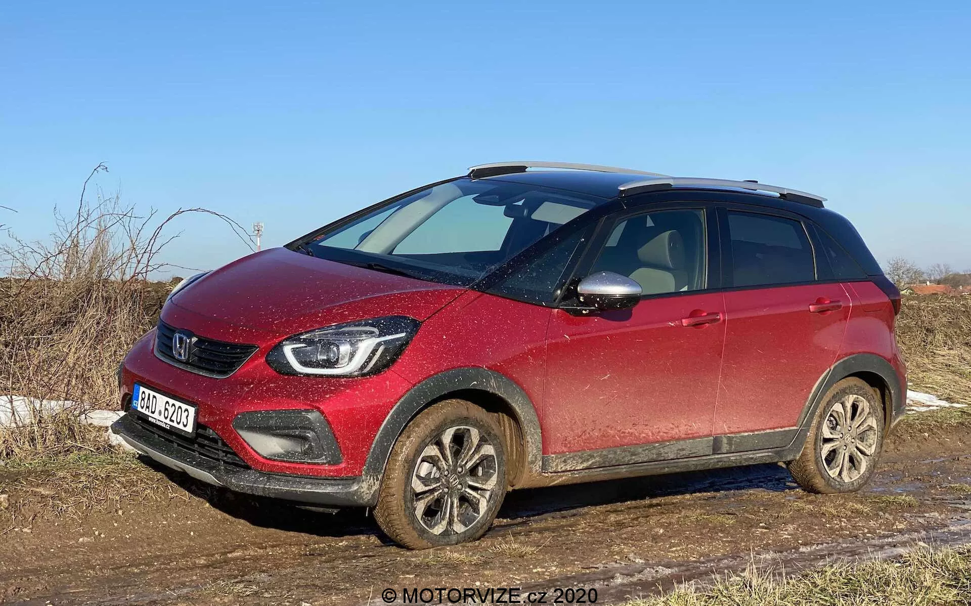 Vue avant trois quarts d'un SUV compact crossover Honda Jazz Crosstar 2020 rouge, capturée à la lumière du jour du côté passager, montrant la calandre avant, les phares, les roues en alliage couvertes de boue et le profil latéral jusqu'à la porte arrière passager. La voiture semble légèrement sale, indiquant une utilisation récente sur des routes non pavées ou mouillées. Les caractéristiques visibles incluent la calandre proéminente avec le logo de la marque, des phares LED modernes, des antibrouillards, des roues en alliage, des rétroviseurs latéraux avec clignotants intégrés, des barres de toit et une garde au sol surélevée, suggérant la polyvalence et la capacité du véhicule pour une utilisation tout-terrain légère. Le SUV est garé dans un cadre rural avec de l'herbe sèche et un ciel dégagé, mettant en avant son adéquation pour les routes de campagne et l'usage tout-terrain léger.