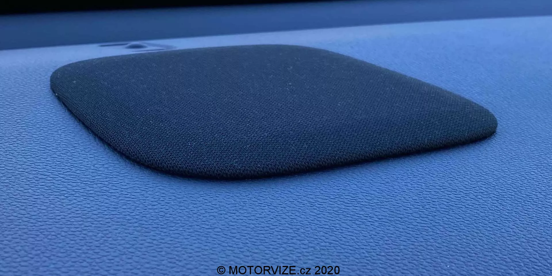 Photographie en gros plan d'une grille de haut-parleur de voiture intégrée dans un tableau de bord ou un panneau de porte de couleur bleu foncé ou noir, peut-être une Honda Jazz 2020. La grille du haut-parleur est fabriquée à partir d'un matériau texturé semblable à du tissu et présente une forme de trapèze tronqué ou de quadrilatère irrégulier avec un côté légèrement convexe. Le composant est légèrement surélevé, projetant une ombre qui met en valeur son profil et sa texture. La photo est prise avec un angle latéral en utilisant une lumière naturelle diffuse, mettant en évidence la texture granuleuse de la surface environnante et contient un filigrane indiquant '© MOTORVIZE.cz 2020.'