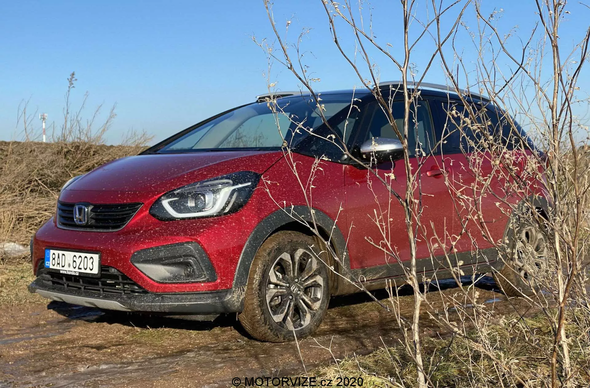 Photo extérieure de jour d'une Honda Jazz 2020 (Jazz Crosstar) stationnée sur un terrain herbeux. L'image capture la couleur rouge vibrante du SUV sous-compact, la calandre proéminente et les optiques de phare modernes avec éclairage LED. La voiture est vue sous un angle de trois quarts avant, montrant sa hauteur de caisse surélevée, ses roues en alliage, son habillage plastique noir autour des arches de roues et des panneaux inférieurs de la carrosserie. Ciel bleu clair, éclairage naturel et branches environnantes qui obscurcissent partiellement la section avant inférieure. Les rétroviseurs avec clignotants intégrés et un aperçu de l'intérieur à travers la fenêtre du conducteur sont visibles.