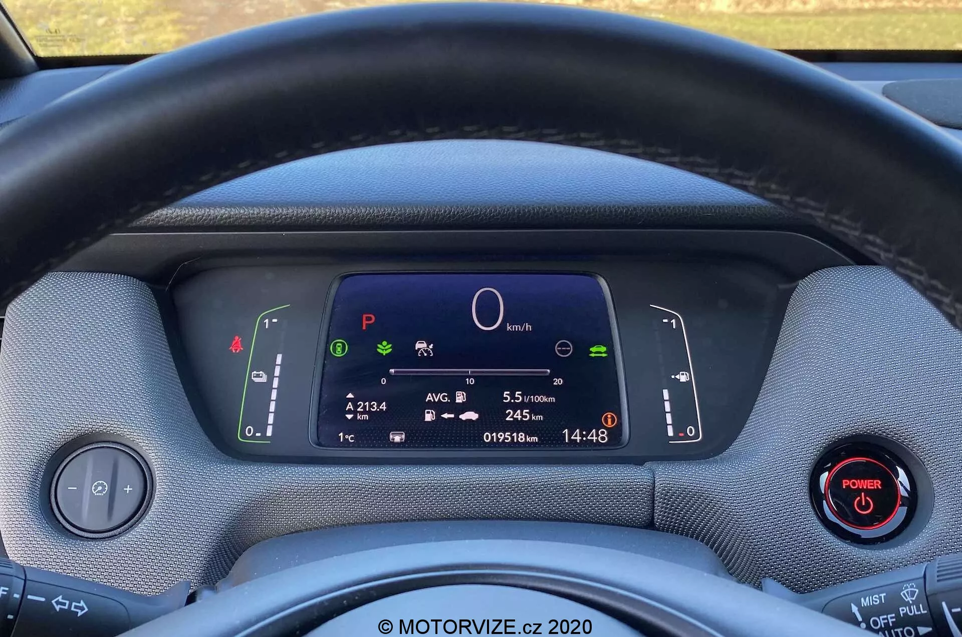 Vue rapprochée de l'intérieur de la Honda Jazz Crosstar 2020 depuis le siège du conducteur, montrant le volant et le tableau de bord entièrement numérique. Le volant habillé de tissu obstrue partiellement la vue de l'affichage numérique, qui présente des symboles de système de voiture, un graphique circulaire central, des informations de trajet, un odomètre et une horloge. Deux manettes derrière le volant contrôlent les clignotants, les feux et les essuie-glaces. Un panneau de commande pour les phares et un bouton de démarrage avec un anneau rouge sont visibles à gauche et à droite, respectivement. Les matériaux du tableau de bord ont l'air texturés, mettant en valeur un design écologique. L'image met en avant la technologie moderne, l'instrumentation numérique et les éventuelles fonctionnalités hybrides ou électriques du véhicule.