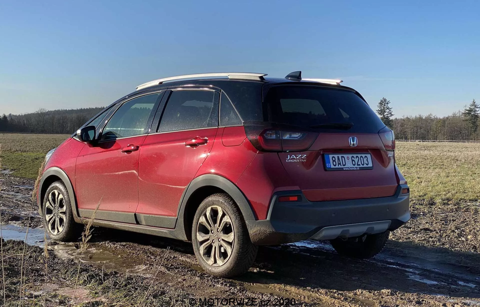 Vue arrière de trois quarts d'une Honda Jazz Crosstar rouge 2020, mettant en avant ses caractéristiques de SUV crossover sous-compact. L'image prise sous un angle bas met en évidence la hauteur et la garde au sol du véhicule, avec des blocs optiques arrière visibles, un badge 'JAZZ', des roues en alliage, un système de barres de toit, des vitres arrière teintées, des éclaboussures de boue et une plaque d'immatriculation visible, garé sur un champ boueux.