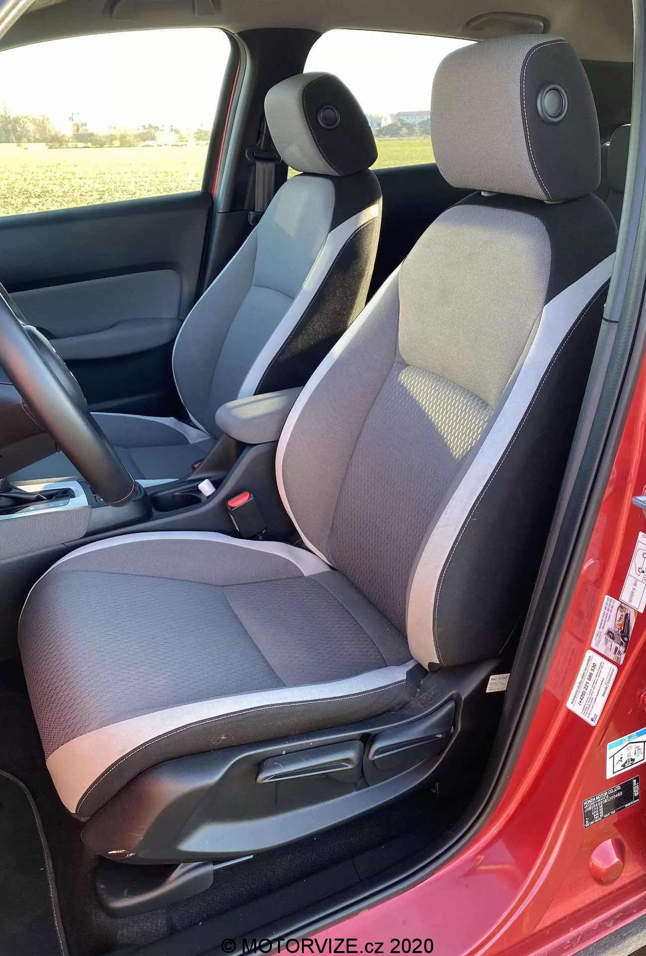Vue intérieure de la Honda Jazz Crosstar 2020 depuis le côté gauche, mettant en avant le siège conducteur en tissu bicolore avec section centrale à motifs, appuie-tête réglable, commandes de réglage manuel du siège, ceinture de sécurité rouge, haut-parleur sur le panneau de porte, tapis de sol sombre, console centrale avec accoudoir, contrôle du rétroviseur latéral et schéma de couleurs sombre et clair.