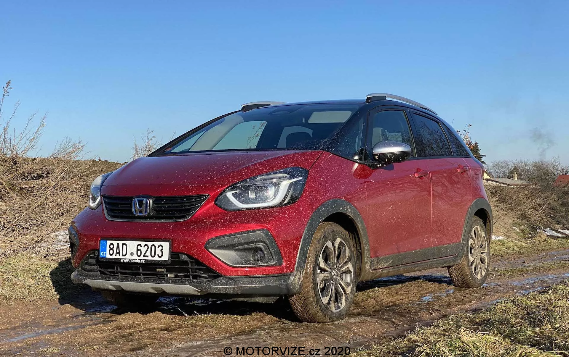 Vue de trois quarts avant d'une Honda Jazz Crosstar 2020 rouge, un SUV crossover compact, capturée sur un terrain tout-terrain avec de la terre et de la boue visibles. L'image met en avant la calandre avant, les phares élégants pouvant loger des lampes LED ou à projecteurs, les feux antibrouillard intégrés au pare-chocs et un capot légèrement incliné. Le véhicule, équipé de barres de toit, de roues couvertes de boue et de rétroviseurs latéraux avec clignotants intégrés, suggère des caractéristiques de sécurité modernes et un équipement technologique avancé. Idéal pour les catégories centrées sur les véhicules, les critiques de voitures, les capacités tout-terrain, la technologie automobile et le design.
