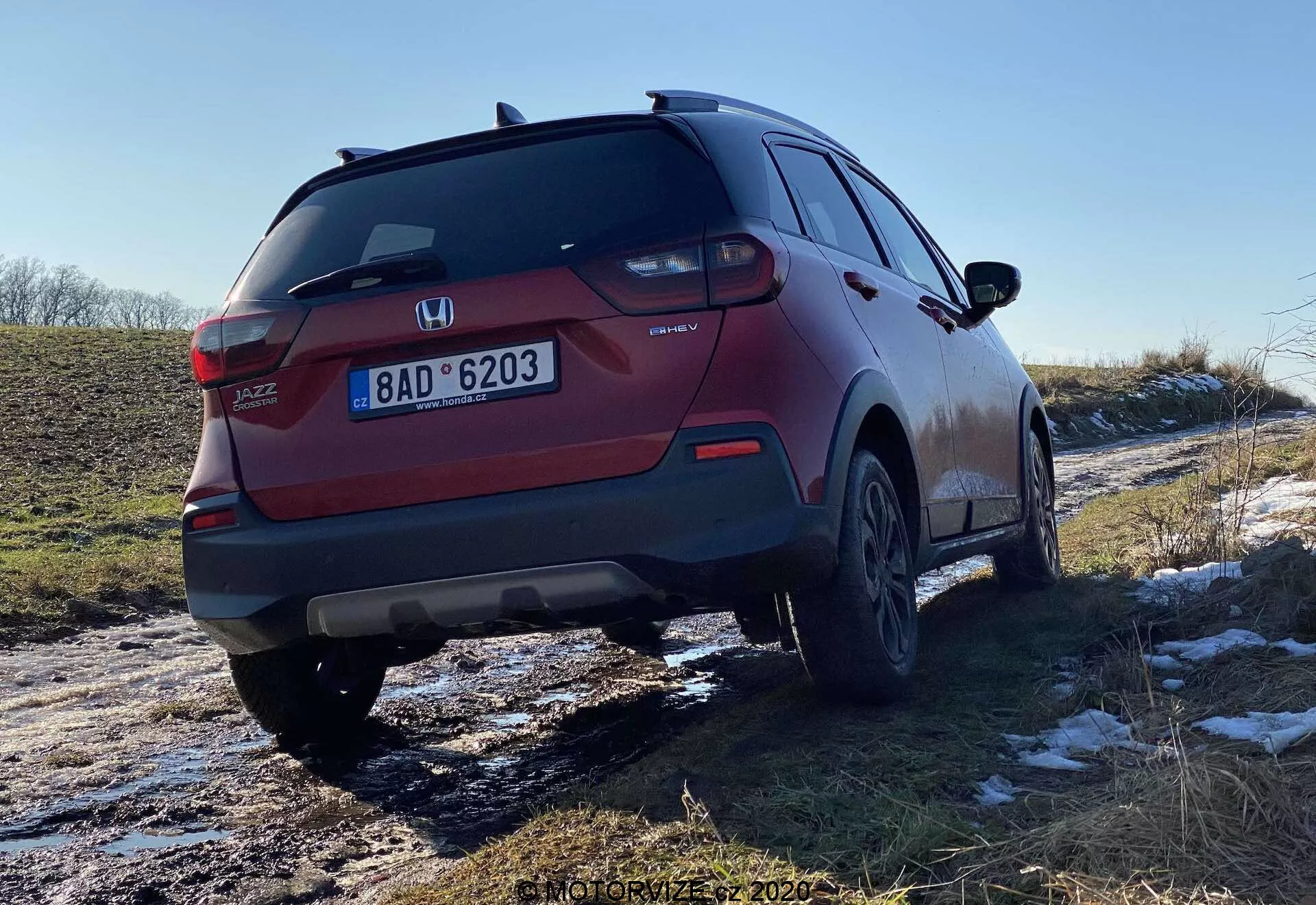 Vue arrière à trois quarts d'un SUV Honda Jazz Crosstar rouge, montrant le design des feux arrière, du pare-chocs arrière, du système d'échappement et des roues en alliage. L'angle met en évidence la garde au sol du véhicule et il semble avoir récemment voyagé sur des routes enneigées et boueuses, indiquant sa polyvalence dans diverses conditions.