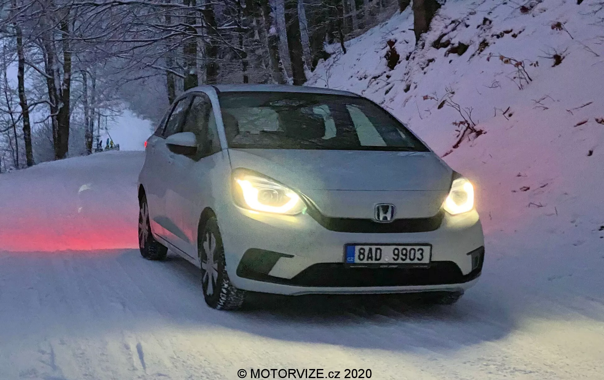 Cette image montre une Honda Jazz 2020, capturée sous un angle de trois quarts avant, mettant en avant les caractéristiques de conception de la calandre avant, des phares et des contours du capot. Positionné sur une route enneigée, le véhicule est équipé de pneus d'hiver avec des motifs de bande de roulement visibles et les phares allumés, indiquant des conditions d'aube, de crépuscule ou de faible visibilité. La plaque d'immatriculation européenne indique « BAD 9903 », et l'image présente un léger flou de mouvement qui ajoute une qualité dynamique. L'éclairage naturel et froid et l'environnement hivernal sont complétés par les capacités tout temps du véhicule.