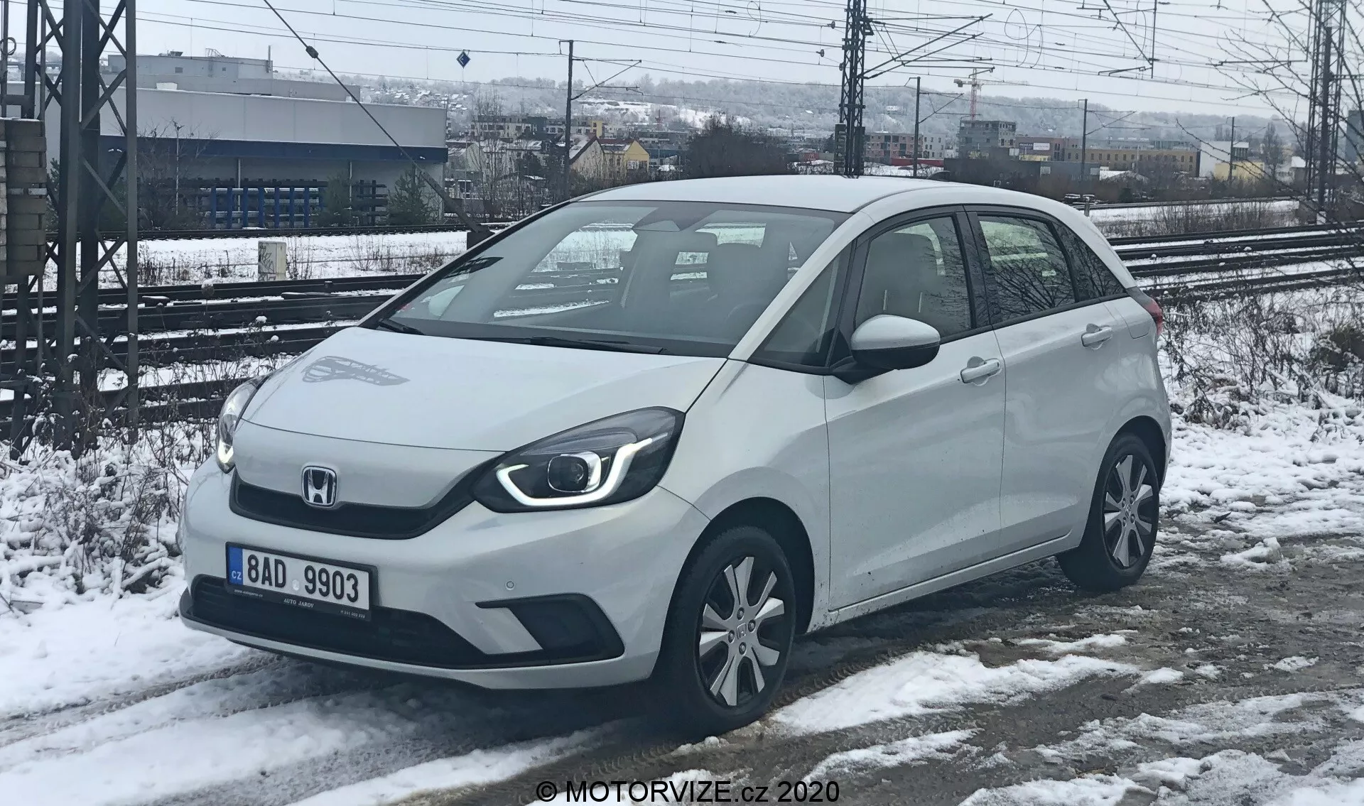 Vue de trois quarts avant d'une voiture compacte Honda Jazz 2020 de couleur claire, possiblement blanche ou argentée, capturée dans un cadre hivernal avec de la neige au sol. Les phares angulaires et la calandre solide suggèrent des options de motorisation électrifiée. La voiture dispose de capteurs de stationnement visibles sur le pare-chocs avant, de rétroviseurs montés sur les portes avec des clignotants intégrés et de jantes en alliage standard. Garée sur de la neige partiellement fondue, avec des voies ferrées et une clôture en arrière-plan, indiquant un emplacement industriel ou proche d'une gare.