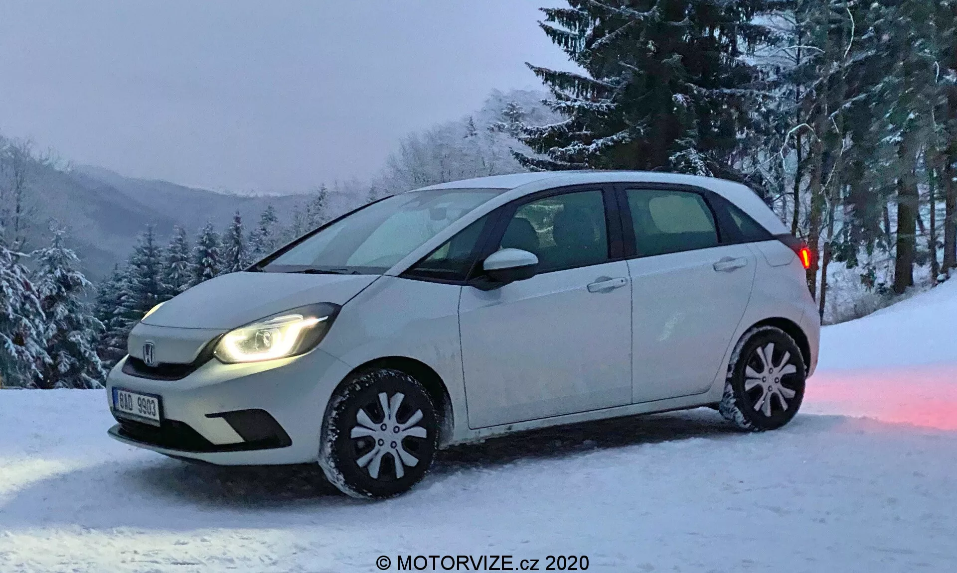 Honda Jazz 2020 hatchback compacte photographiée sous un angle frontal oblique capturant une vue complète du côté conducteur et une vue partielle du pare-chocs avant et de la calandre. Le véhicule présente une combinaison de couleurs bicolores avec la majorité en couleur claire et la partie inférieure des portes latérales dans une teinte plus sombre. Les phares sont allumés, avec des lampes halogènes typiques, sans feux antibrouillard. Les enjoliveurs de roue d'hiver décoratifs sont visibles avec des flocons de neige réfléchissants, indiquant une saison hivernale. La voiture est garée sur une zone enneigée avec un fond d'arbres et une colline enneigée, suggérant un lieu rural ou montagneux. Image courtoisie de MOTORVIZE.cz.