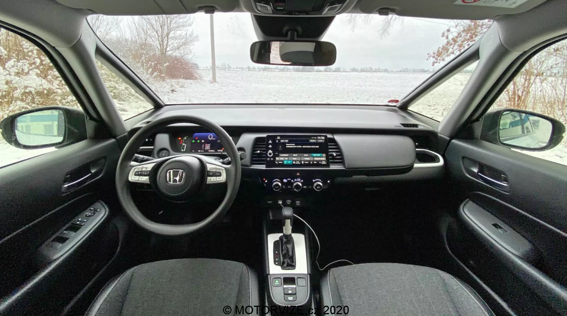 Intérieur de voiture moderne vu depuis la banquette arrière, comprenant un tableau de bord numérique, un volant multifonctionnel, un grand écran tactile d'infodivertissement, une boîte de vitesses automatique avec levier en cuir, une climatisation à double zone, un frein de parking électronique et un design élégant avec des matériaux contrastants; extérieur enneigé visible à travers le pare-brise, indiquant une possible ambiance hivernale.