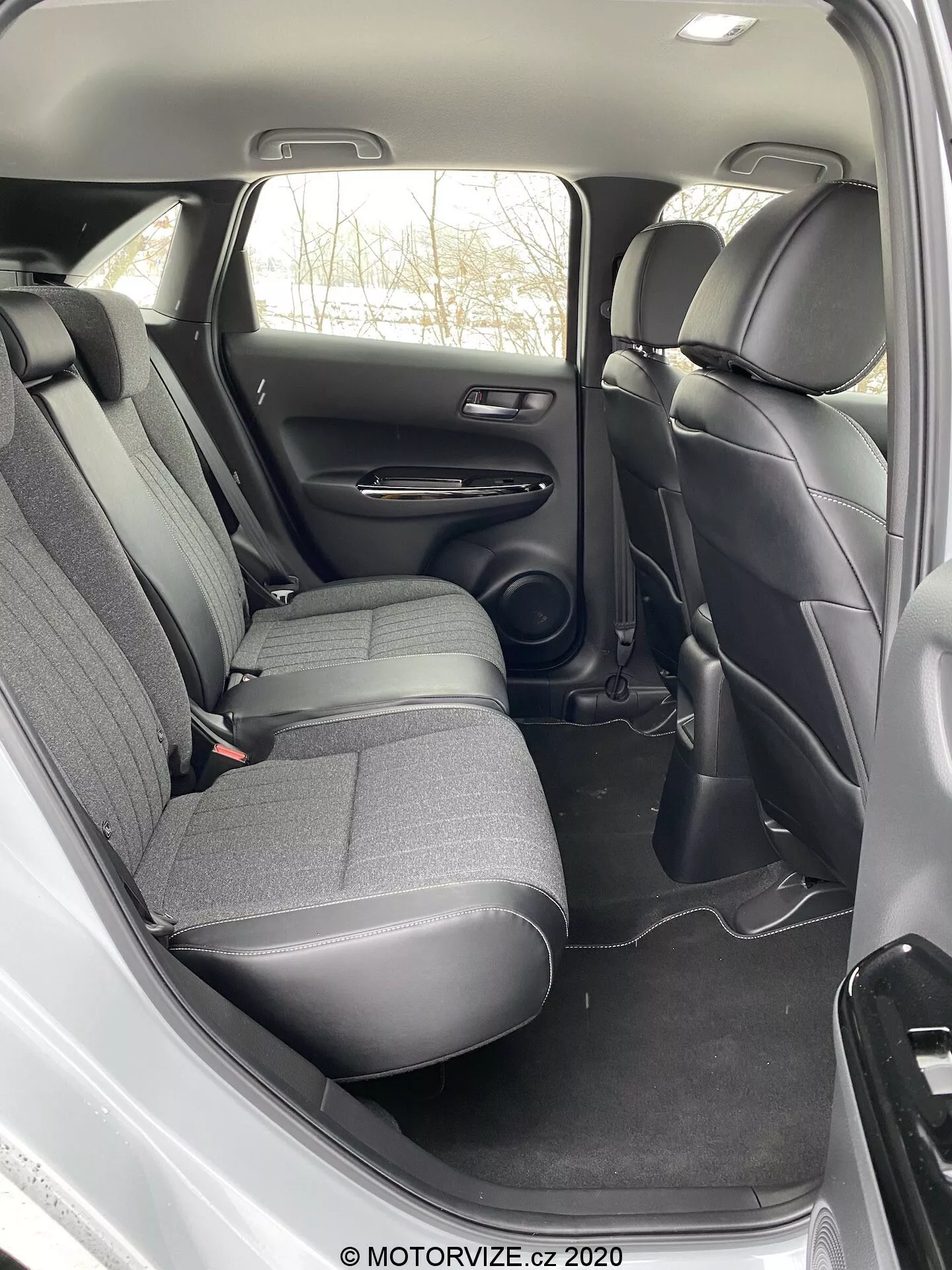 L'image montre l'habitacle arrière d'une Honda Jazz 2020. La vue, prise d'un angle légèrement en hauteur au-dessus et à côté de la porte arrière ouverte, révèle une banquette arrière avec appuie-têtes pour trois passagers, revêtue d'un tissu durable de couleur sombre. Le panneau de porte comporte des commandes, un haut-parleur, un accoudoir et un compartiment de rangement, tandis que le sol est recouvert d'un tapis de teinte sombre assortie. Entre les sièges avant, une console centrale s'étend vers l'arrière, sans contrôle climatique arrière visible ni points de charge. Les dispositifs de sécurité visibles comprennent des ceintures de sécurité à trois points. Le design est pratique avec des matériaux standards, mettant l'accent sur la durabilité et la fonctionnalité plutôt que sur le luxe.
