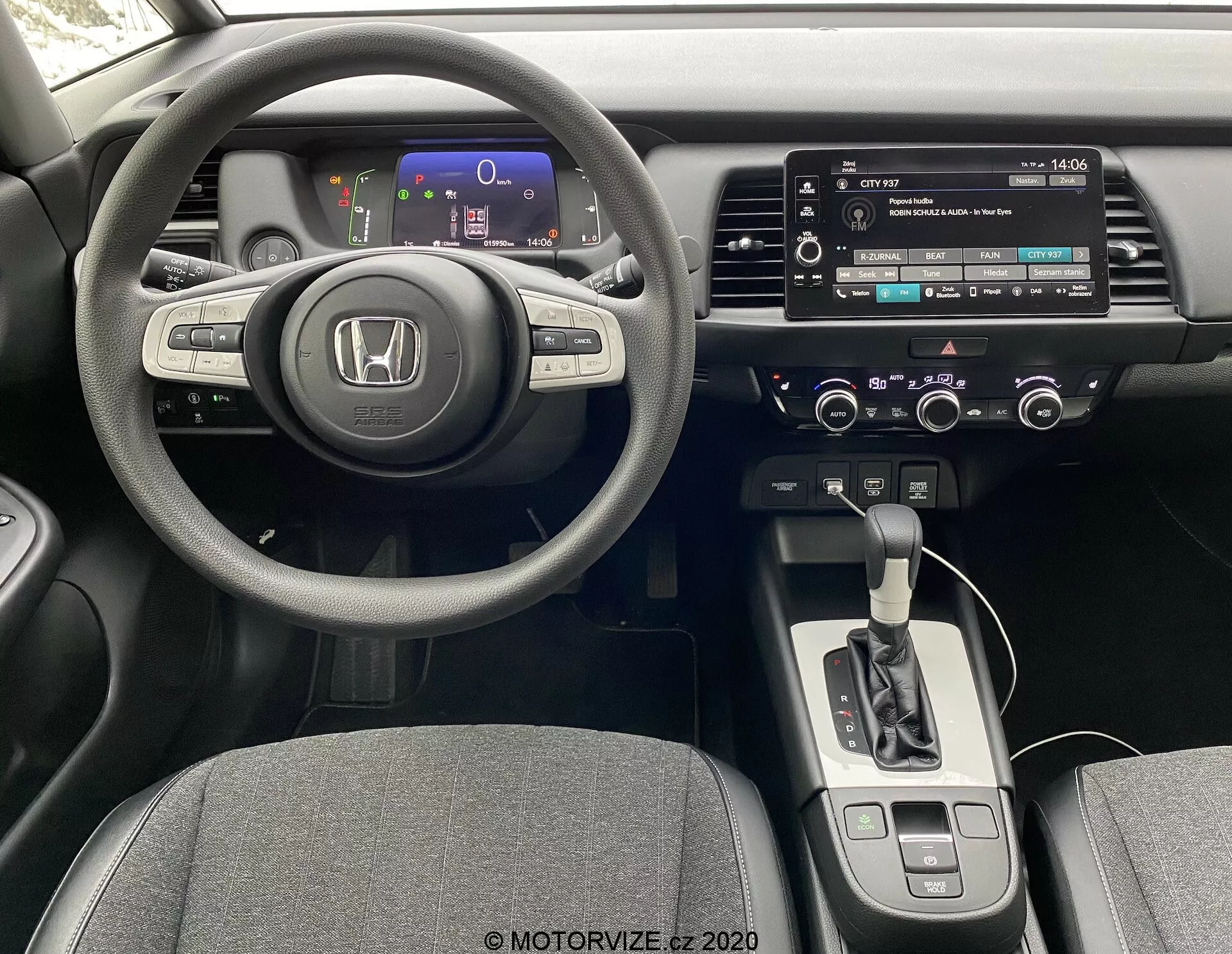 Intérieur d'une Honda Jazz 2020 vu de la perspective du conducteur, montrant un volant à trois branches avec commandes intégrées, un tableau de bord numérique avec des jauges colorées, une console centrale avec système d'infodivertissement à écran tactile, des commandes manuelles pour l'audio et le climat, un sélecteur de transmission automatique, et des sièges en tissu.
