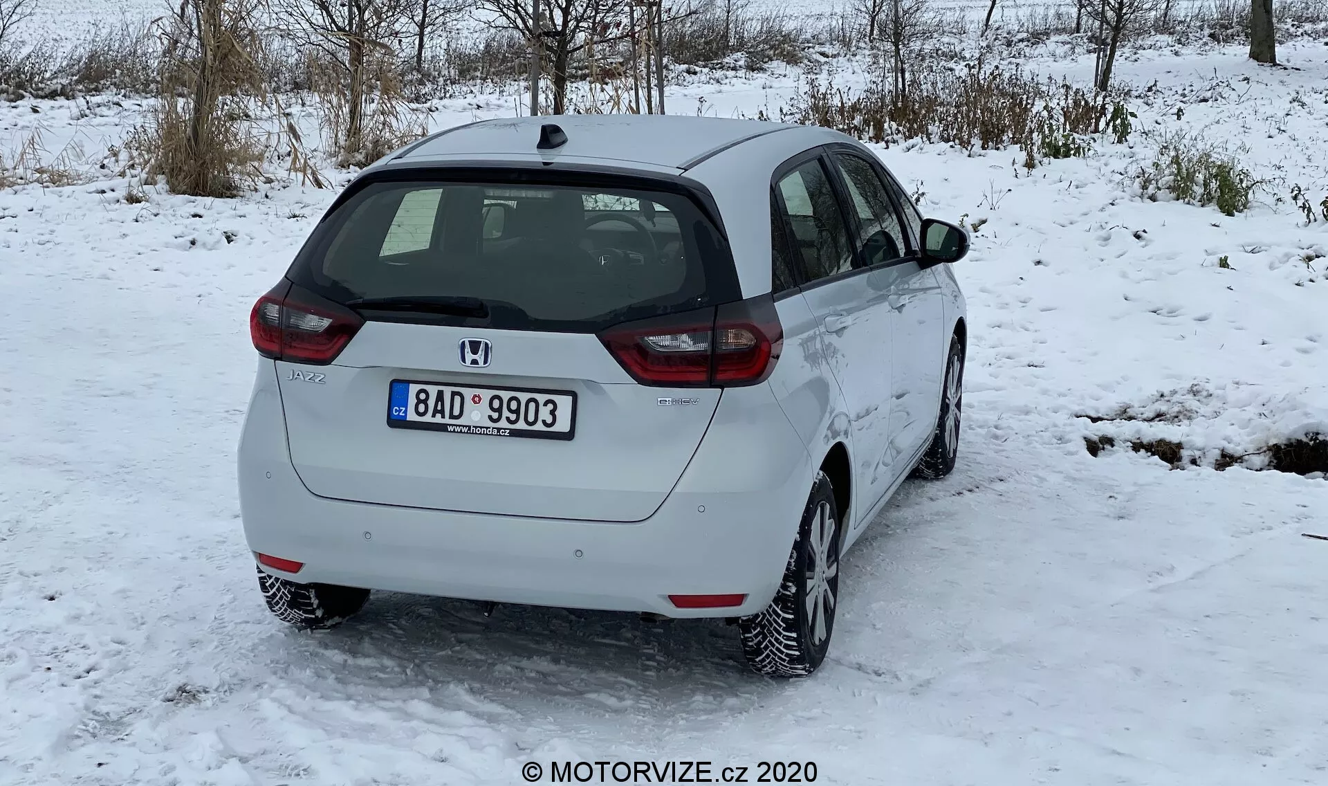 L'image montre une vue de trois quarts arrière d'une Honda Jazz 2020 blanche sur une surface enneigée, avec des arbres sans feuilles dans un décor hivernal et un ciel couvert en arrière-plan. Les caractéristiques du design arrière incluent un essuie-glace arrière, l'emblème du fabricant, l'indication du modèle et l'assemblage des feux arrière avec des lentilles rouges et transparentes. Le profil latéral montre la porte arrière passager, la porte avant, les fenêtres latérales et un bouchon de carburant visible. La voiture est équipée de jantes en alliage à plusieurs rayons, indiquant une bande de roulement des pneus adaptée à la neige avec une légère accumulation de neige. La plaque d'immatriculation arrière suggère son origine géographique. La garde au sol est typique des hatchbacks, sans embout d'échappement visible ni accessoires extérieurs. La voiture semble propre et en bon état, avec peu de saleté sur le pare-chocs arrière en raison des routes enneigées.