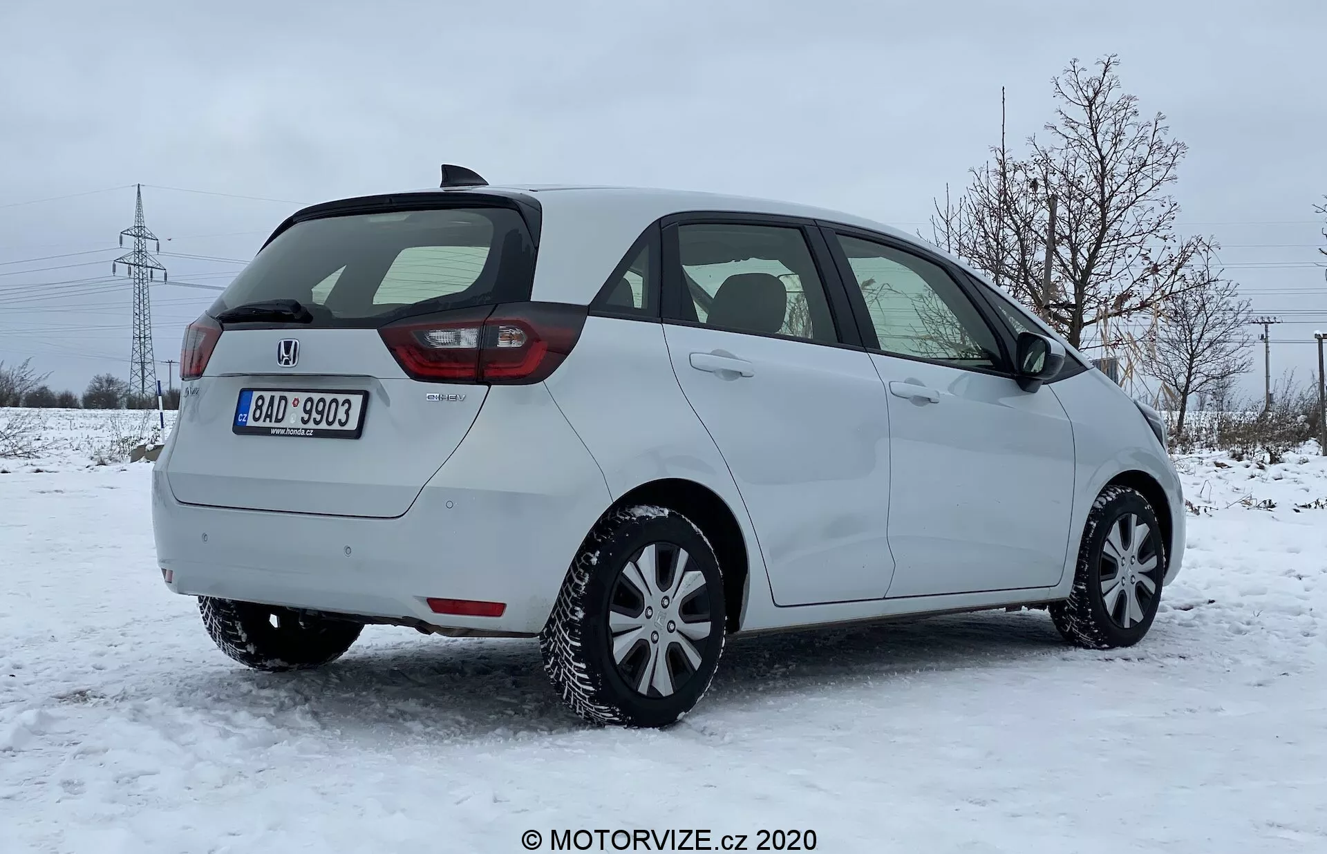 Vue arrière trois quarts d'une voiture hatchback Honda Jazz 2020 sur une surface couverte de neige dans un cadre suburbain avec des arbres dénudés et des pylônes électriques en arrière-plan. L'image capture le côté passager de la voiture, le design moderne des feux arrière probablement avec une technologie LED, le hayon arrière, l'essuie-glace arrière, le pare-chocs arrière visible avec des capteurs de stationnement intégrés et probablement des pneus d'hiver. Le véhicule est stationné en plein jour par un ciel couvert, suggérant des conditions de conduite hivernales.