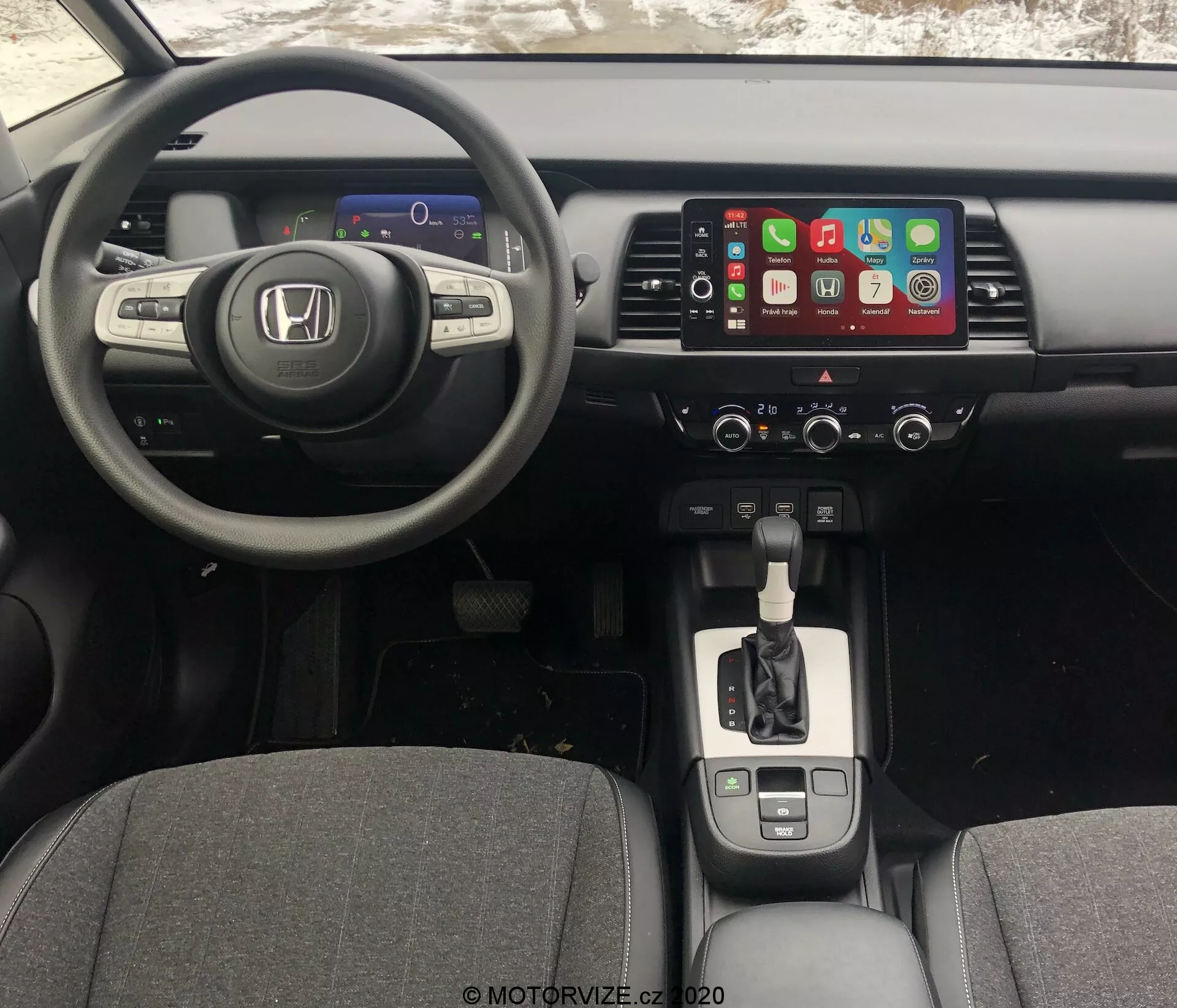 Vue intérieure de la Honda Jazz 2020 depuis la perspective du conducteur, montrant le tableau de bord, le volant et la console centrale. L'angle légèrement élevé révèle le tableau de bord numérique, le volant multifonctionnel à trois branches, le système d'info-divertissement à écran tactile avec Apple CarPlay, les commandes de climatisation, le levier de vitesse avec finition chromée, les porte-gobelets, le frein de stationnement électronique et la connectivité USB.