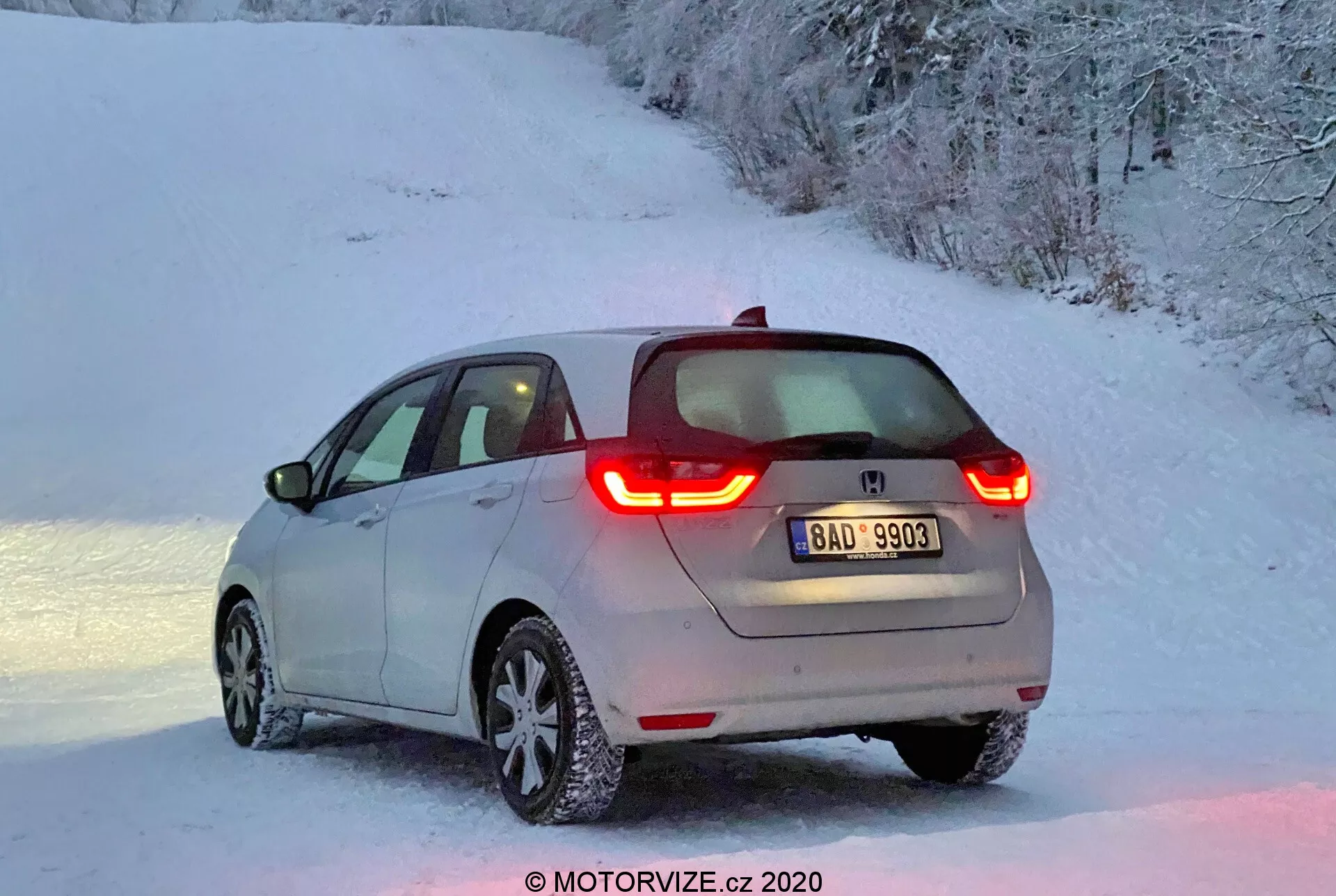 Vue arrière trois quarts d'une Honda Jazz 2020 moderne, éclairée par la douce lumière de l'aube ou du crépuscule, avec des feux arrière à LED en forme de C allumés, un logo du fabricant visible, une légère couche de neige sur le véhicule et des jantes en alliage avec des pneus couverts de neige dans un cadre rural enneigé.
