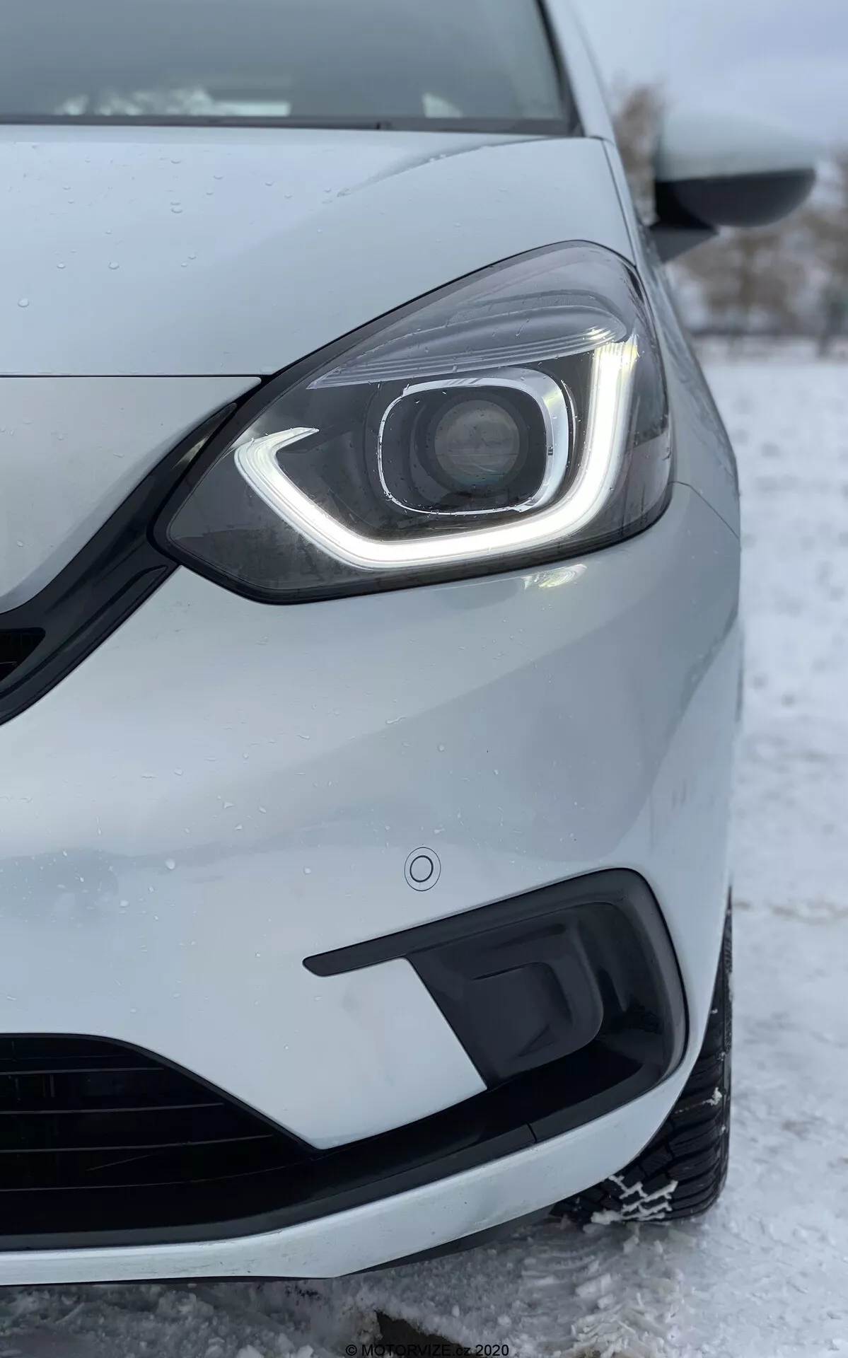 Gros plan de la partie avant gauche d'une Honda Jazz 2020, pris en contre-plongée. L'accent est mis sur le phare qui présente un design moderne à LED avec des feux de jour courbant autour du haut et du côté. L'assemblage du phare comprend une lentille de projecteur visible, suggérant des phares HID ou LED avancés pour le faisceau principal. Le pare-chocs avant, montré sous le phare, inclut une grille d'admission d'air et un logement pour les phares antibrouillard. La peinture métallique argenté ou gris clair de la voiture et la roue partiellement visible et le rétroviseur latéral sont détaillés. La neige et la glace au sol indiquent un climat froid, reflétant les performances de la voiture dans des conditions hivernales. L'image met également en évidence la qualité de fabrication de la voiture, les écarts entre les panneaux et le capteur de proximité sur le pare-chocs, qui font partie de la technologie d'assistance au stationnement.