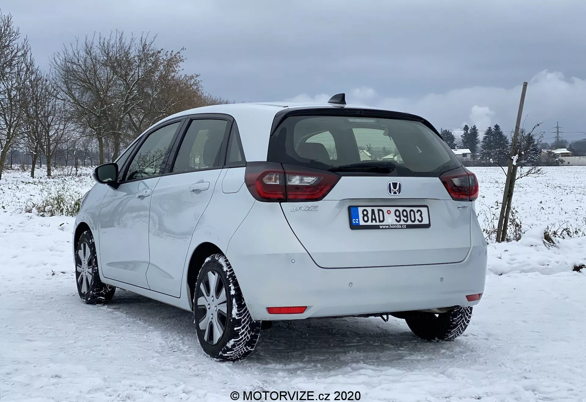Image d'une Honda Jazz 2020 argentée garée dans des conditions enneigées avec un paysage rural ou semi-rural en arrière-plan. Capturée depuis l'arrière et de profil partiel droit, mettant en évidence les feux arrière disposés verticalement, le badge central et le pare-chocs avec capteurs de stationnement. Les contours latéraux lisses, les jantes en alliage et les pneus d'hiver sont clairement visibles, ainsi que la capacité de dégivrage des vitres et du pare-brise arrière, et les rétroviseurs latéraux non rabattables de la même couleur que la carrosserie. La plaque d'immatriculation, l'antenne de toit et l'état général bien entretenu du véhicule sont également notés.