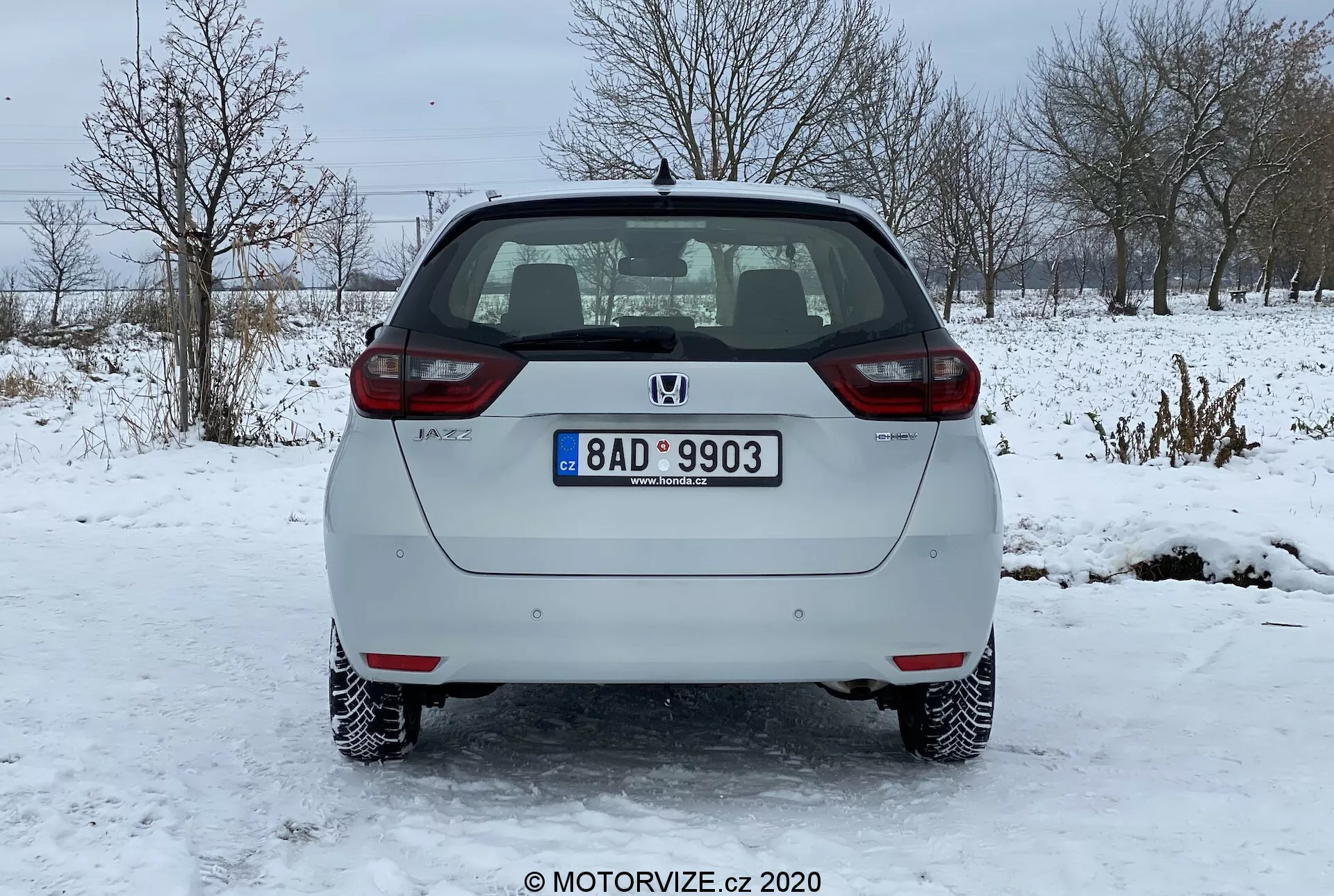 Vue arrière d'une voiture hatchback argentée Honda Jazz 2020 garée sur une surface glacée. L'image capture clairement le pare-chocs arrière, le hayon, les feux arrière et le pare-brise arrière du véhicule. Le logo du fabricant et le nom du modèle sont visibles sur le hayon, ainsi qu'une plaque d'immatriculation standard montée au centre. La voiture est équipée de lumières de plaque d'immatriculation illuminées, de réflecteurs aux deux extrémités du pare-chocs arrière et de feux arrière à ampoules standards. Le pare-brise arrière possède une fonction de dégivrage indiquée par des lignes horizontales, et un essuie-glace arrière est stationné en haut. Le système d'échappement est légèrement visible sous le pare-chocs arrière du côté droit. La voiture a des roues en acier avec des enjoliveurs décoratifs et se trouve dans un paysage hivernal avec de la neige et des arbres nus en arrière-plan.