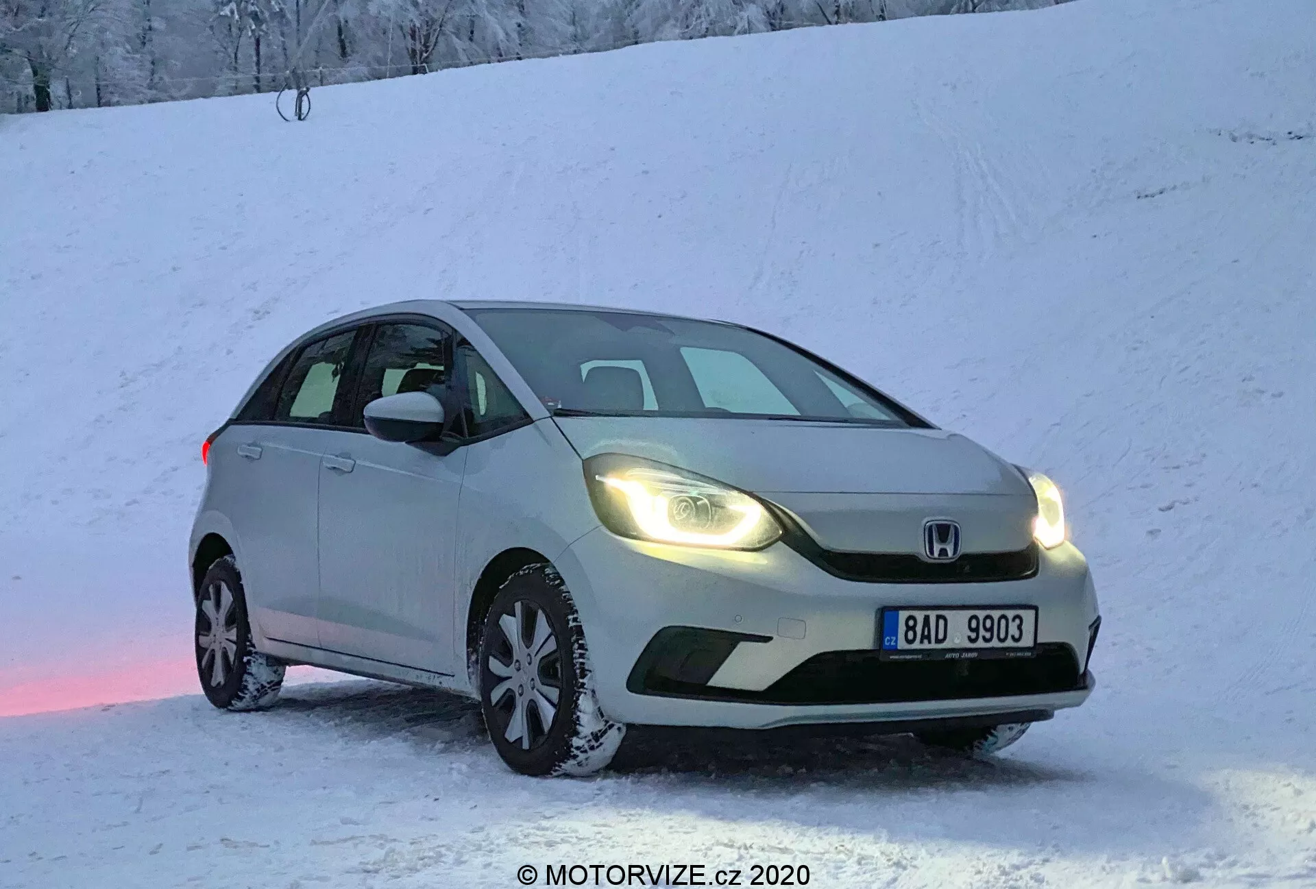Voiture Honda Jazz 2020 de couleur argent garée sur une surface enneigée au lever ou au coucher du soleil, avec les phares allumés, capturée en vue trois quarts avant. L'image met en évidence la calandre avant, les phares, le capot, le pare-brise et une partie du côté conducteur. Plaque d'immatriculation européenne visible, neige sur le sol et la voiture, créant une ambiance froide mais chaleureuse. Légère réflexion de lumière rose sur la neige sous la voiture. Prise de vue en angle bas qui met en valeur l'équipement des pneus d'hiver et l'aspect dynamique pour les capacités de conduite hivernales.