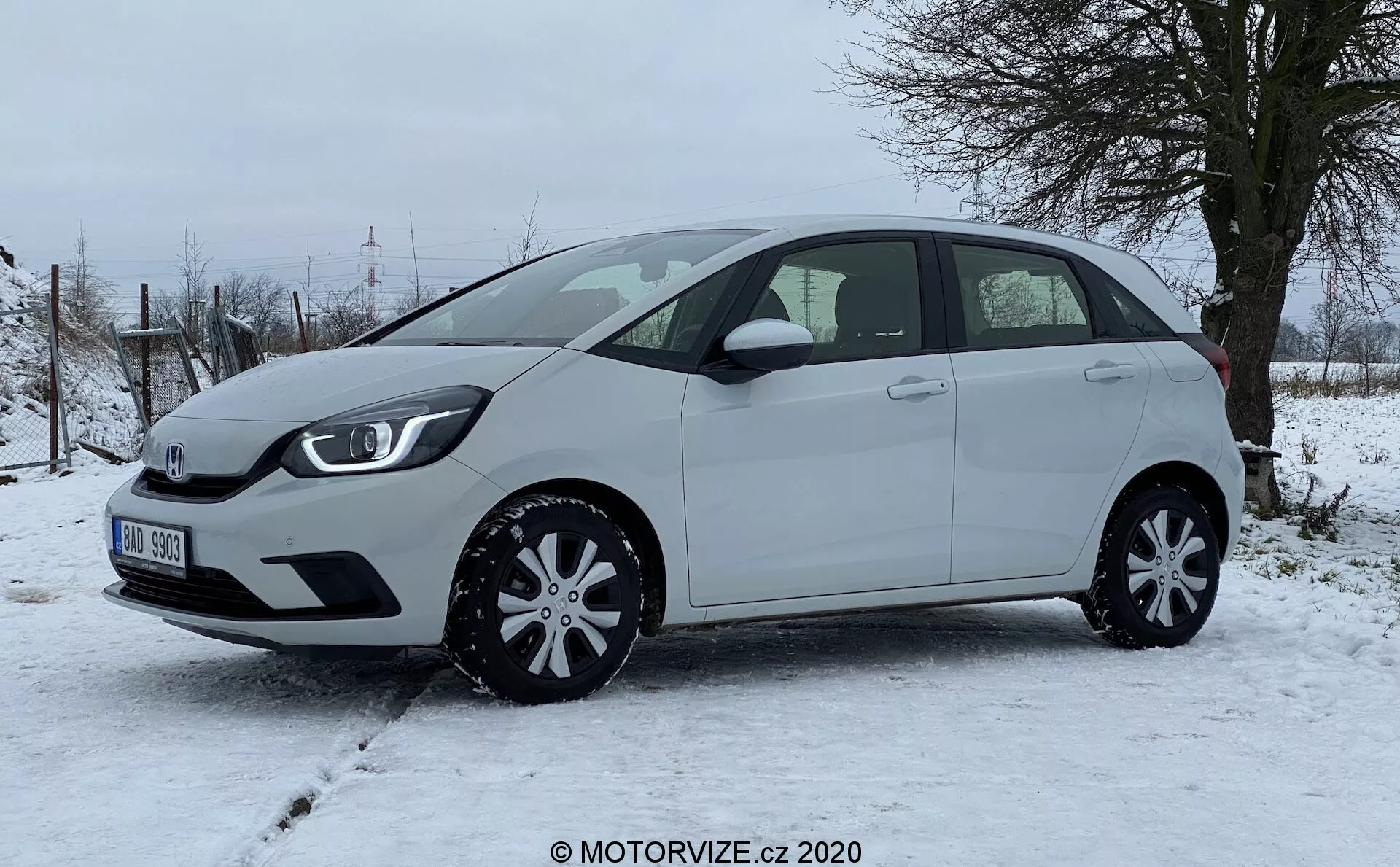 Un moderne Honda Jazz 2020 hatchback photographié de côté, mettant en valeur le profil gauche du véhicule sur un terrain enneigé avec une végétation rurale en arrière-plan. La voiture compacte présente une ligne de toit légèrement inclinée, cinq portes dont une arrière, des rétroviseurs latéraux de couleur carrosserie et des enjoliveurs élégants, démontrant la praticité et l'efficacité du design. Aucune caractéristique extérieure avancée comme des caméras ou des capteurs n'est visible sous cet angle, soulignant une vue claire des proportions, des contours des portes, de la ligne des fenêtres et des passages de roues du véhicule.